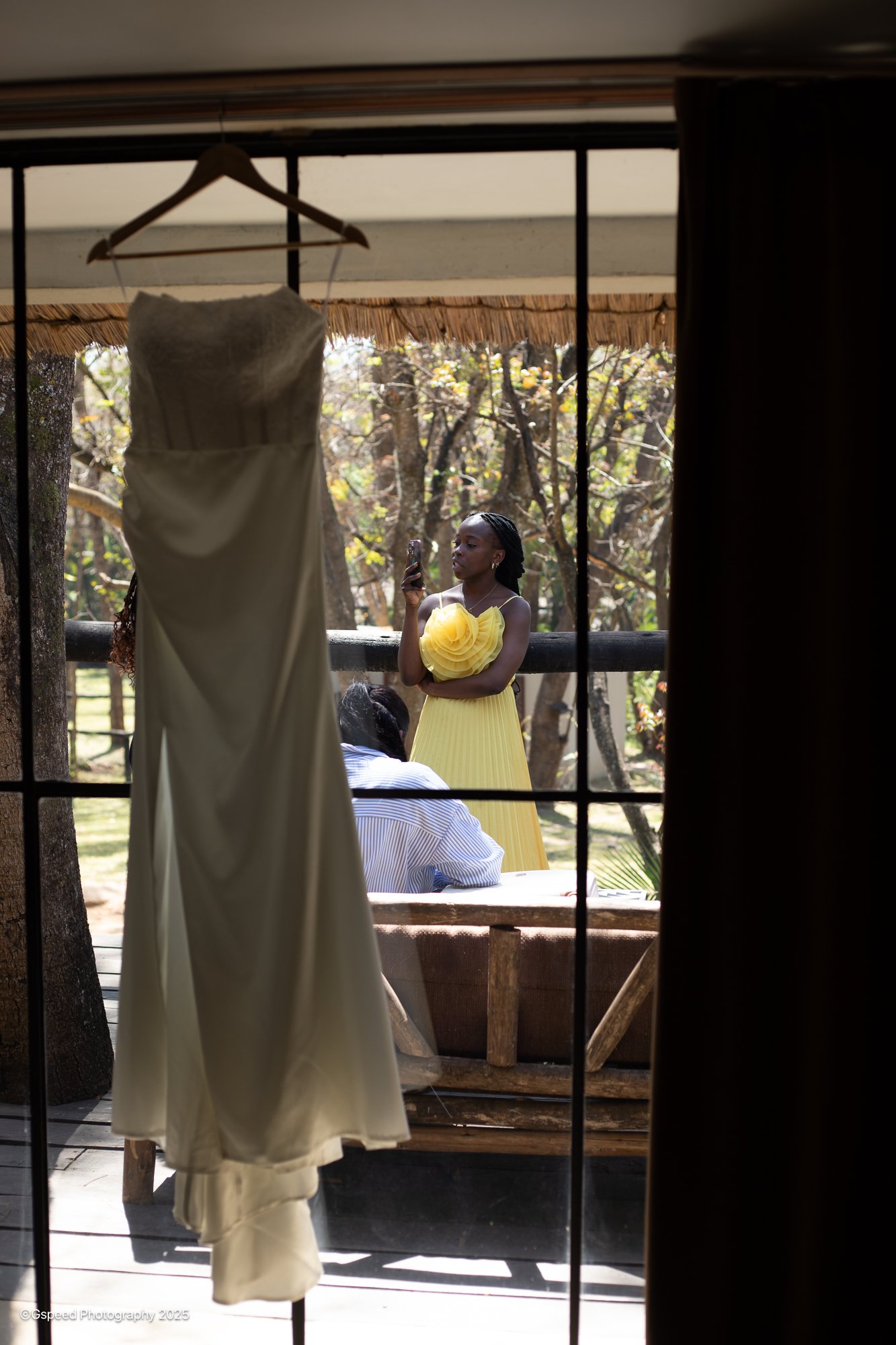 A wedding dress hanging on a hanger inside a house, seen through a window with black metal bars. Outside, a woman in a yellow dress is holding a smartphone, standing in an outdoor area with trees in the background.