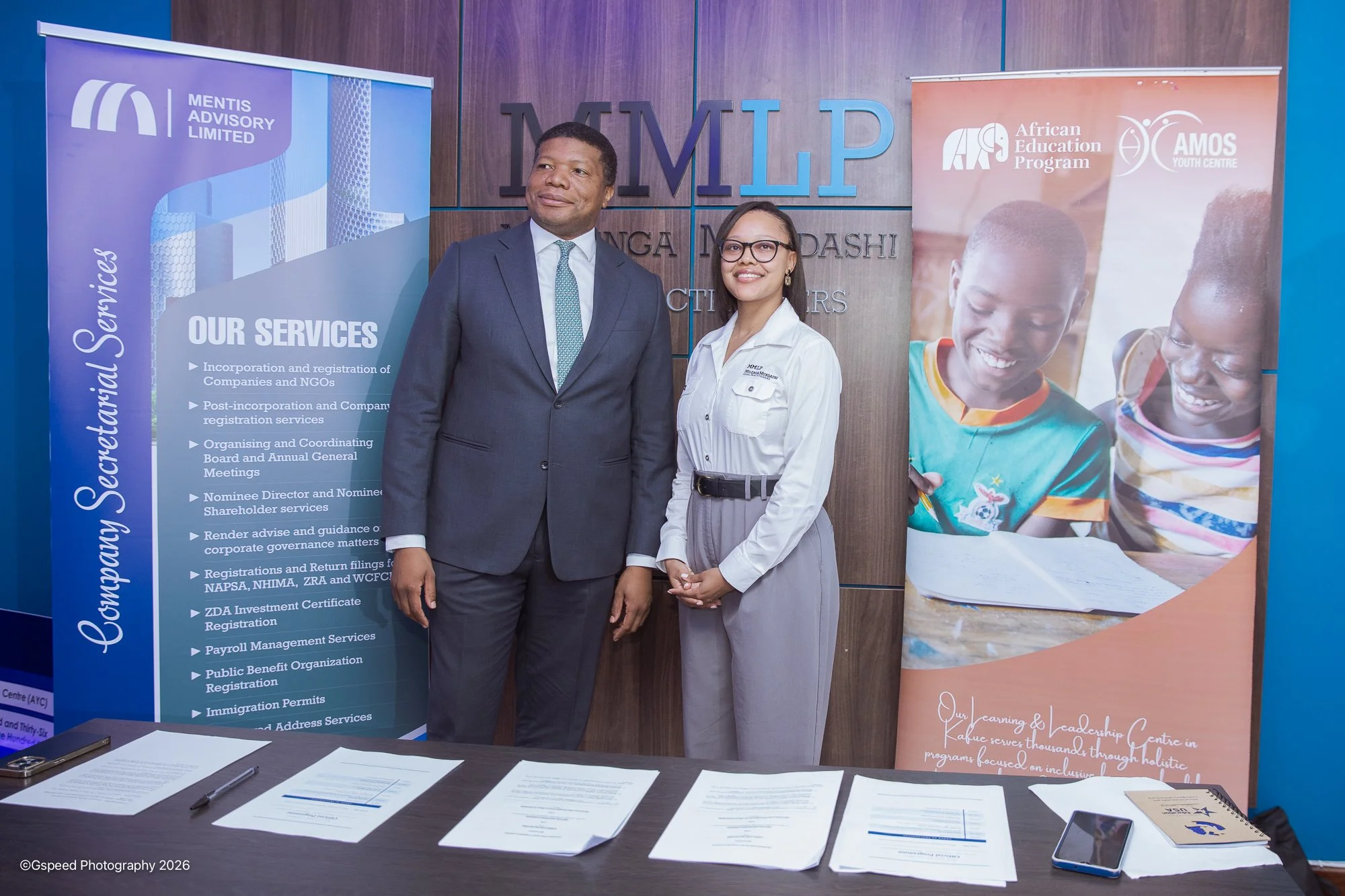 A man in a suit and a woman in glasses and a white shirt standing in front of banners at a conference or event. The banners display organization names and services related to African Education Program and Mentoring Leadership Project.