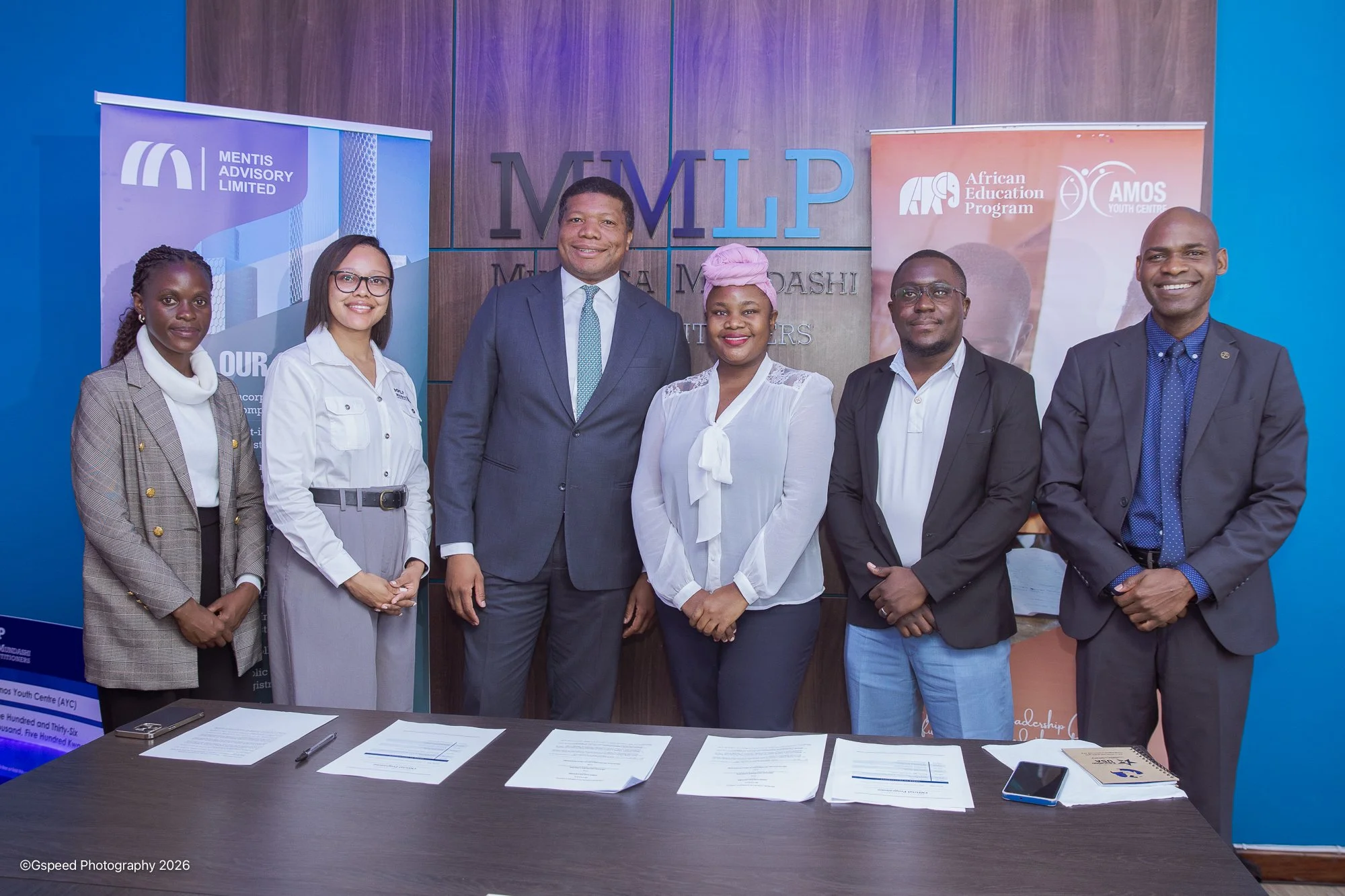 Group of six people standing behind a table with documents, in front of a wooden wall with corporate logos and banners for Mentis Advisory Limited, African Education Program, and AMOS Youth Centre, during a professional event.