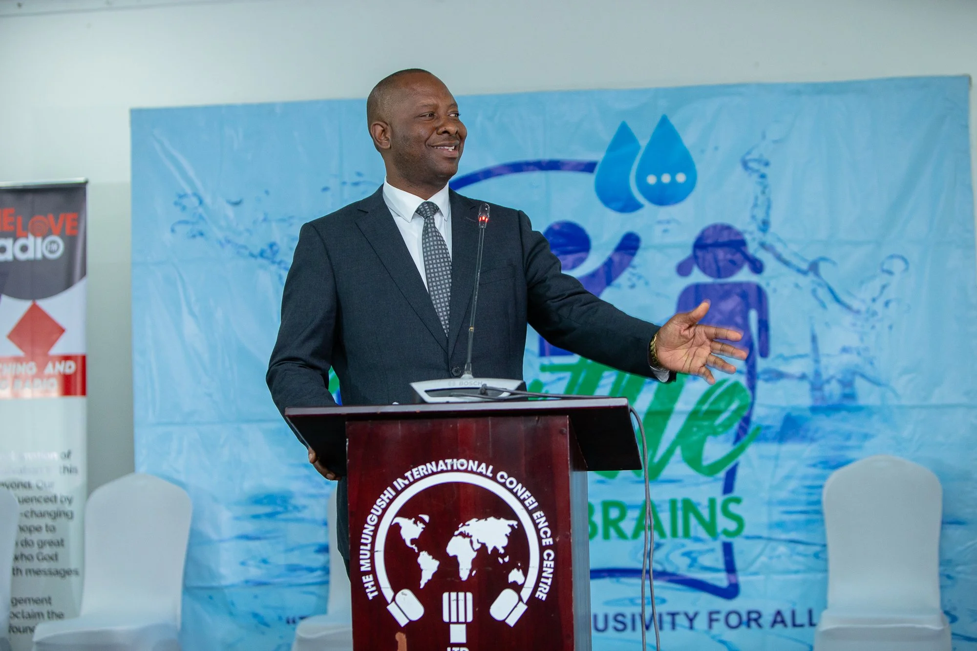 A man in a suit and tie standing at a podium delivering a speech or presentation. Behind him is a blue banner with a water droplet and insect logo, and a partial banner on the side with the words 'LOVE radio' visible.