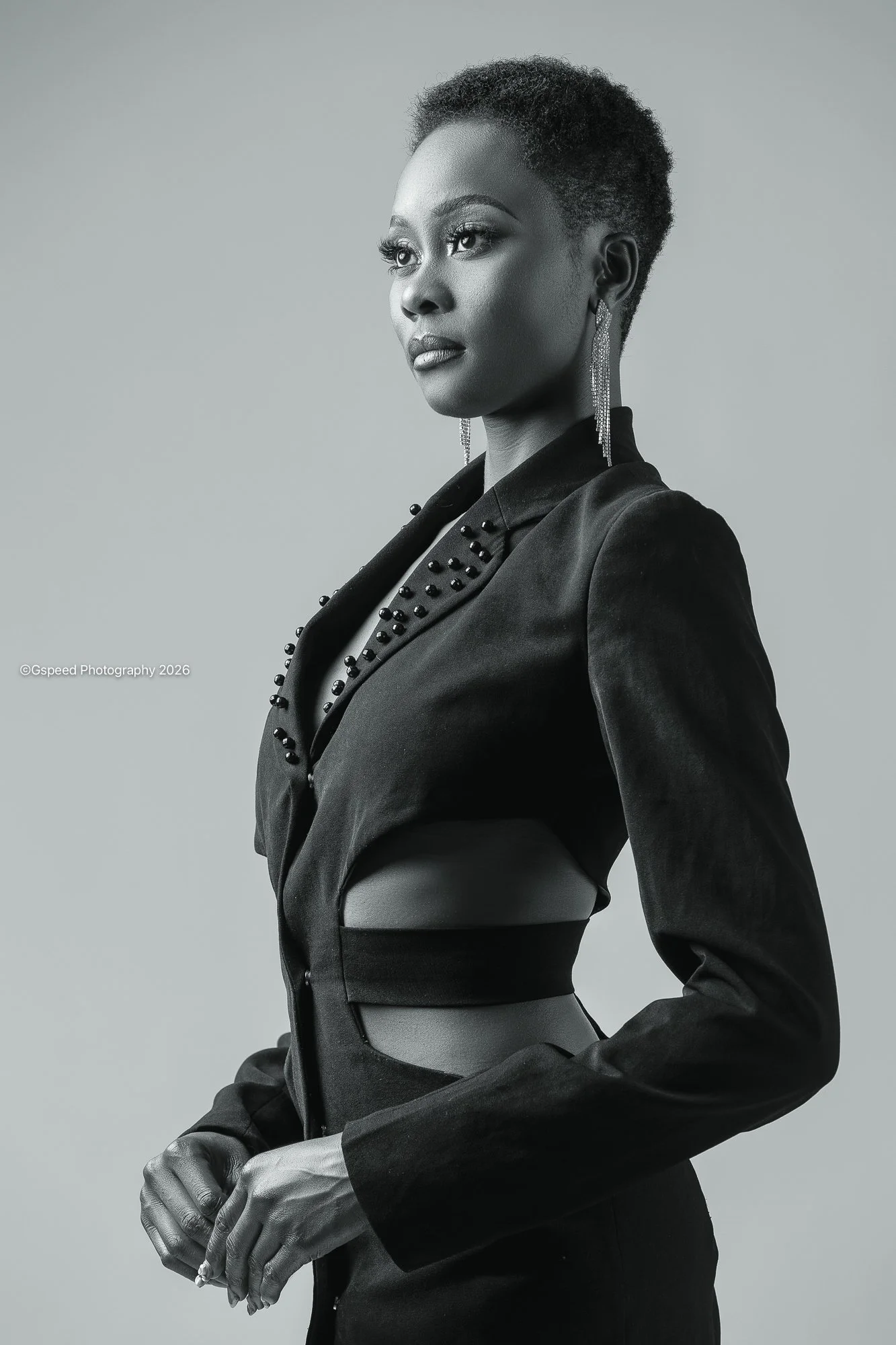 Black woman with short curly hair in a black jacket with bead embellishments, wearing long earrings, posed with hands clasped, in a gray studio background.