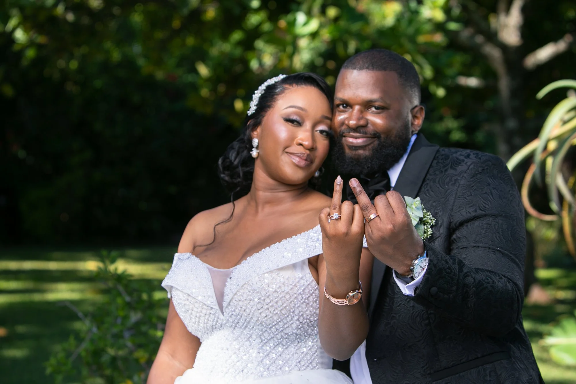 A newly married couple posing outdoors, standing close with their foreheads touching and hands showing wedding rings. The bride is wearing a white wedding dress with beaded details and a pearl headband, while the groom is dressed in a black tuxedo wi