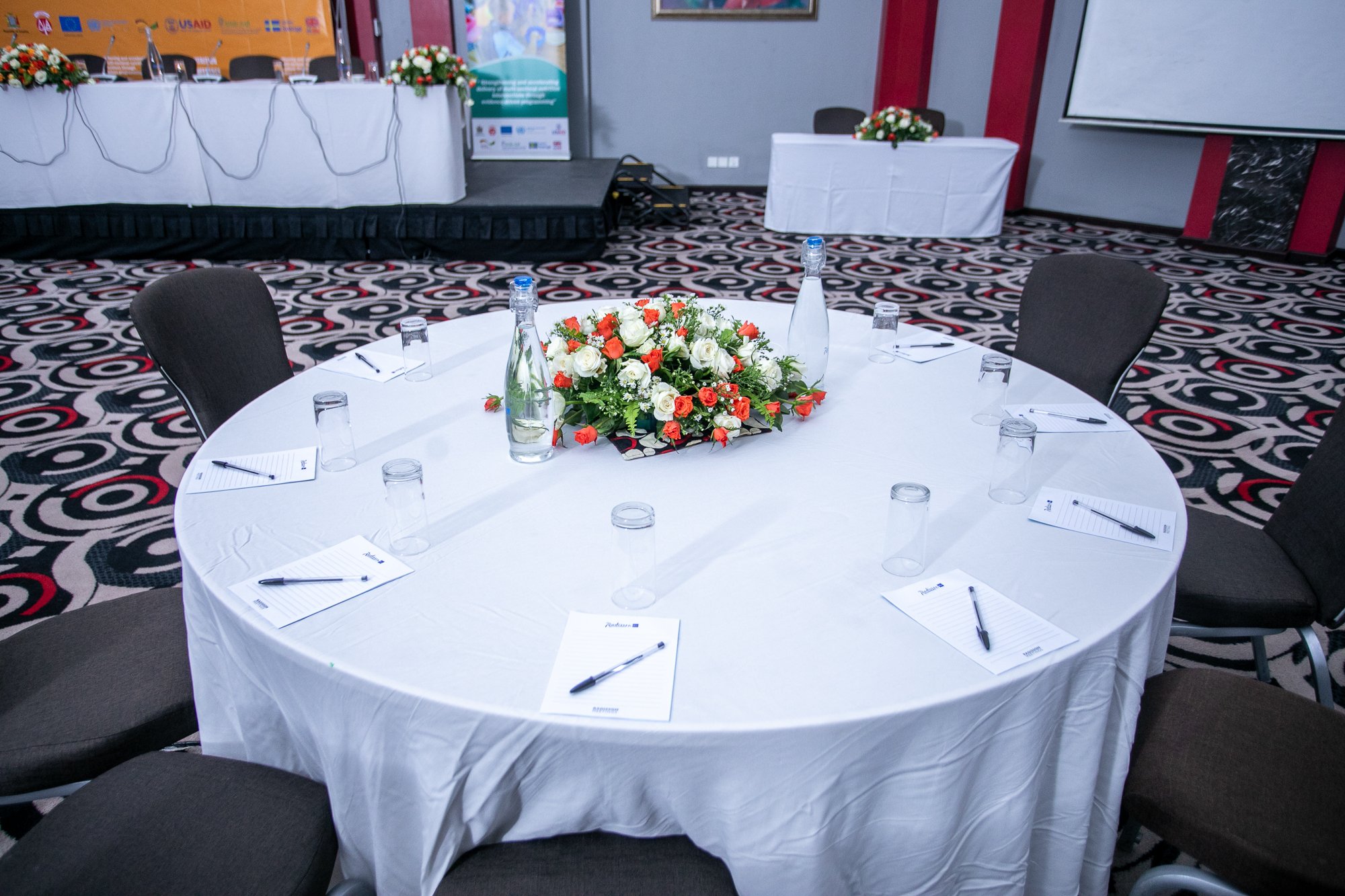 A conference room set up for a formal meeting or event, with a round table covered in a white tablecloth, decorated with a large floral centerpiece of white, red, and orange flowers. Around the table are six black chairs, each with notepads and pens,