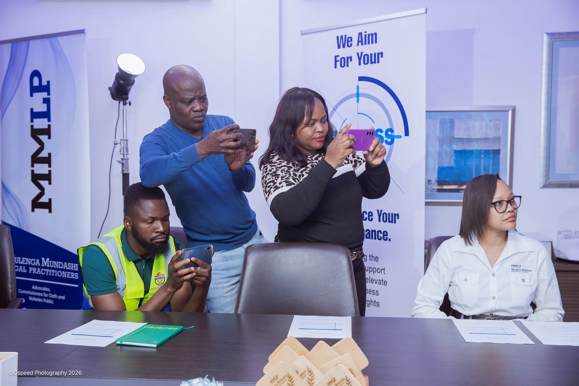 Group of people in a meeting room, some taking photos with their phones, with banners and framed pictures on the wall.