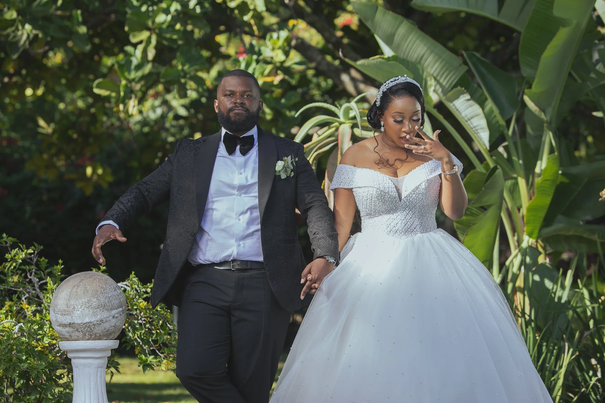 A bride and groom holding hands outdoors among green foliage, with the bride wearing a white dress and tiara, and the groom in a black tuxedo with a bow tie, during a wedding or special occasion.