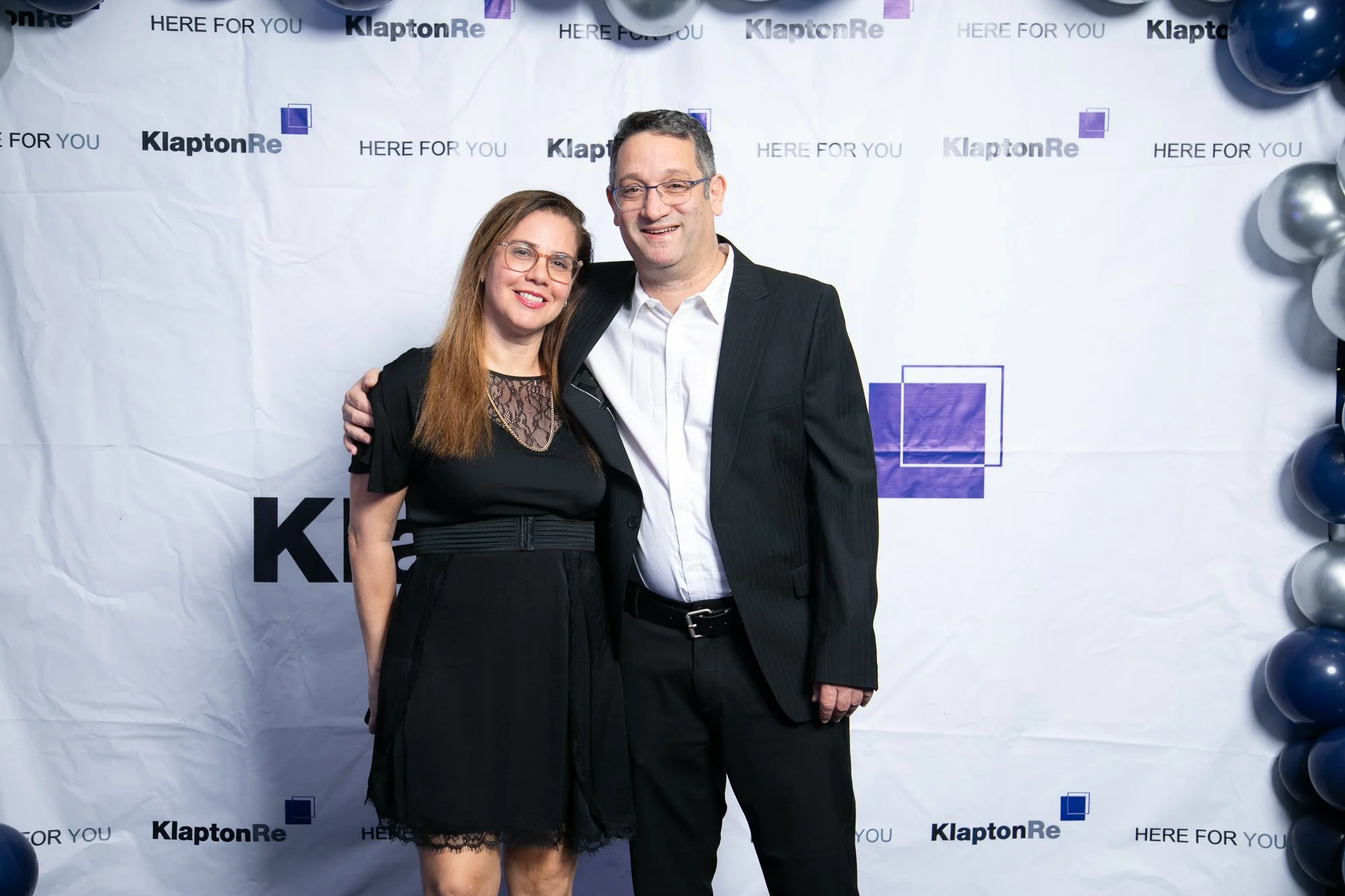 A man and woman smiling and standing close together at a photo booth with a backdrop that reads "KlaptonRe" and "HERE FOR YOU," surrounded by balloons.