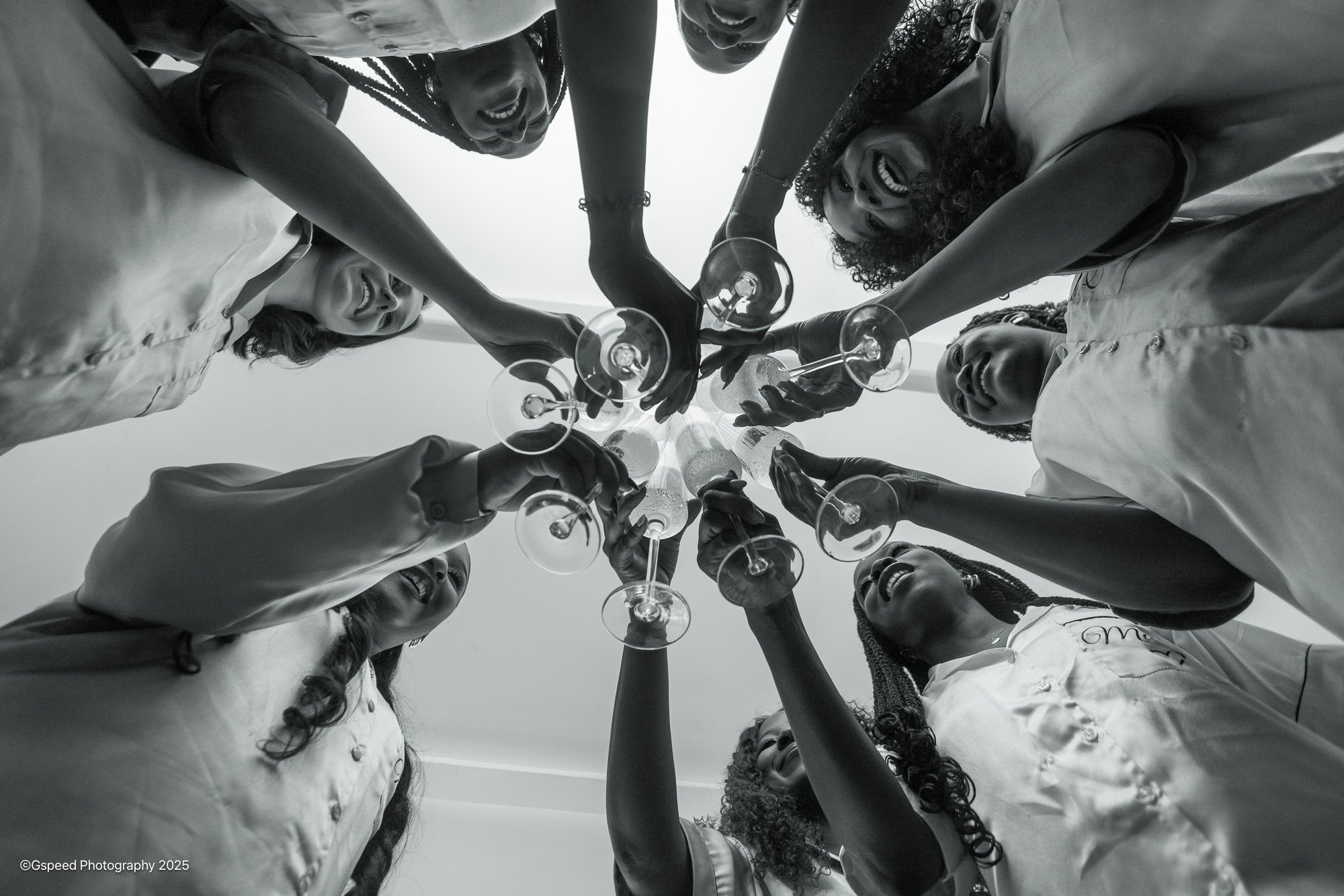 A group of diverse women are standing in a circle, laughing and smiling, holding glasses of wine, taken from a low-angle perspective.