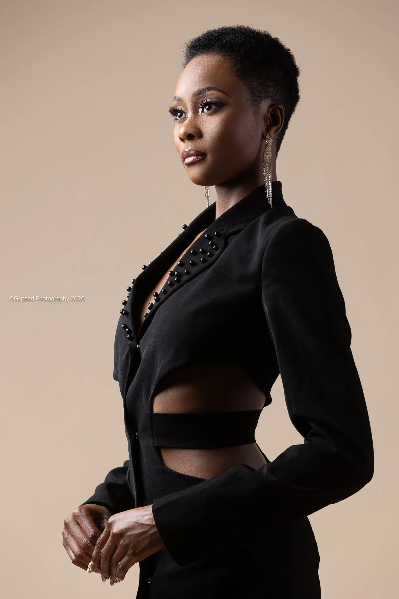 A Black woman with short curly hair wearing a black blazer with bead embellishments, long dangling earrings, and black pants against a beige background.