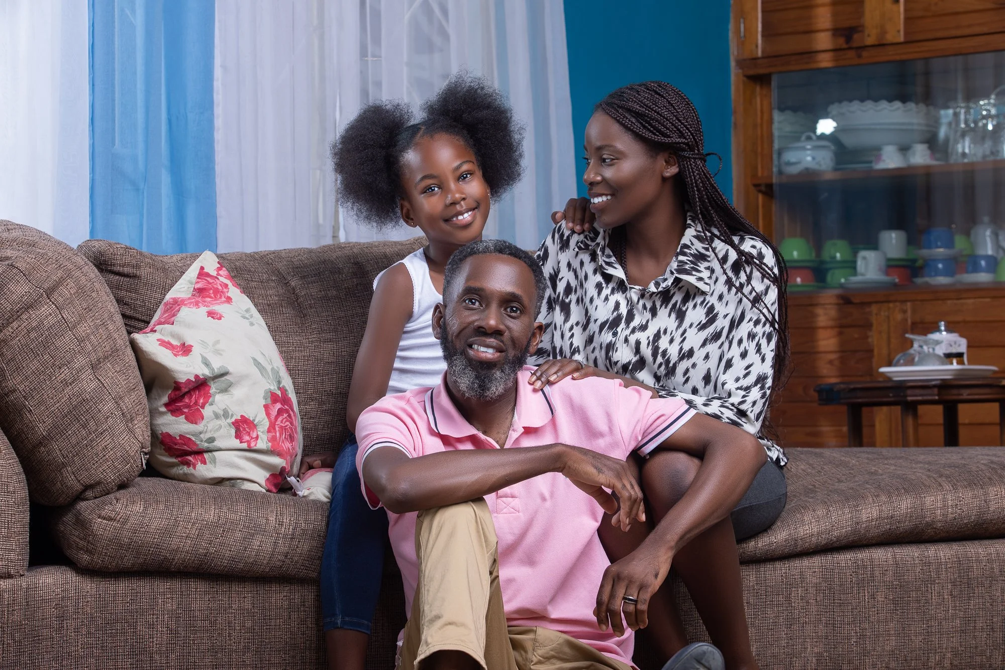 Family of three sitting on a sofa in a living room, smiling and interacting with each other. There is a decorative pillow on the sofa, and a wooden cabinet with dishes in the background.