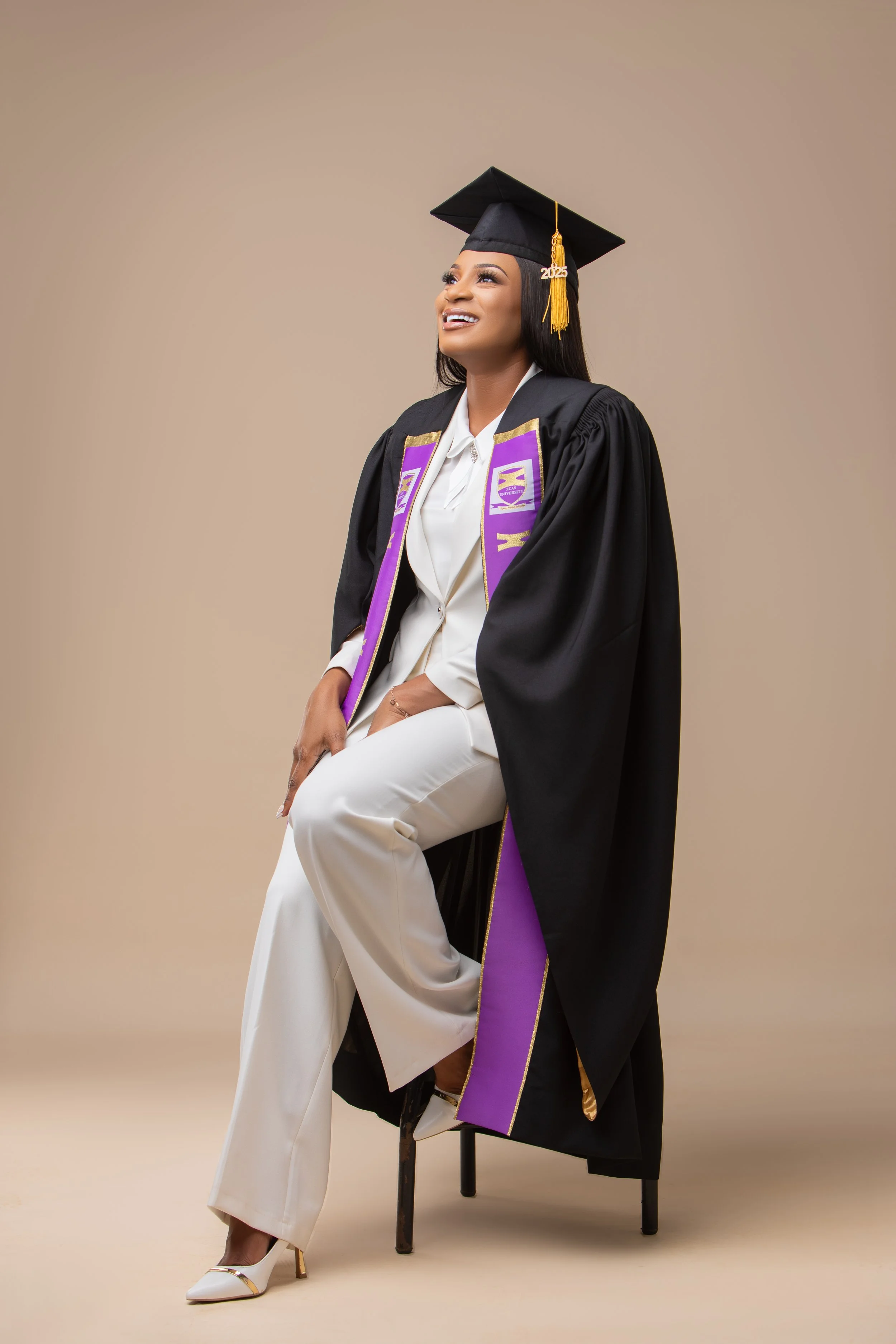 A woman in a graduation gown and cap with a '2025' tassel is sitting on a chair, smiling and looking upward, against a beige background.