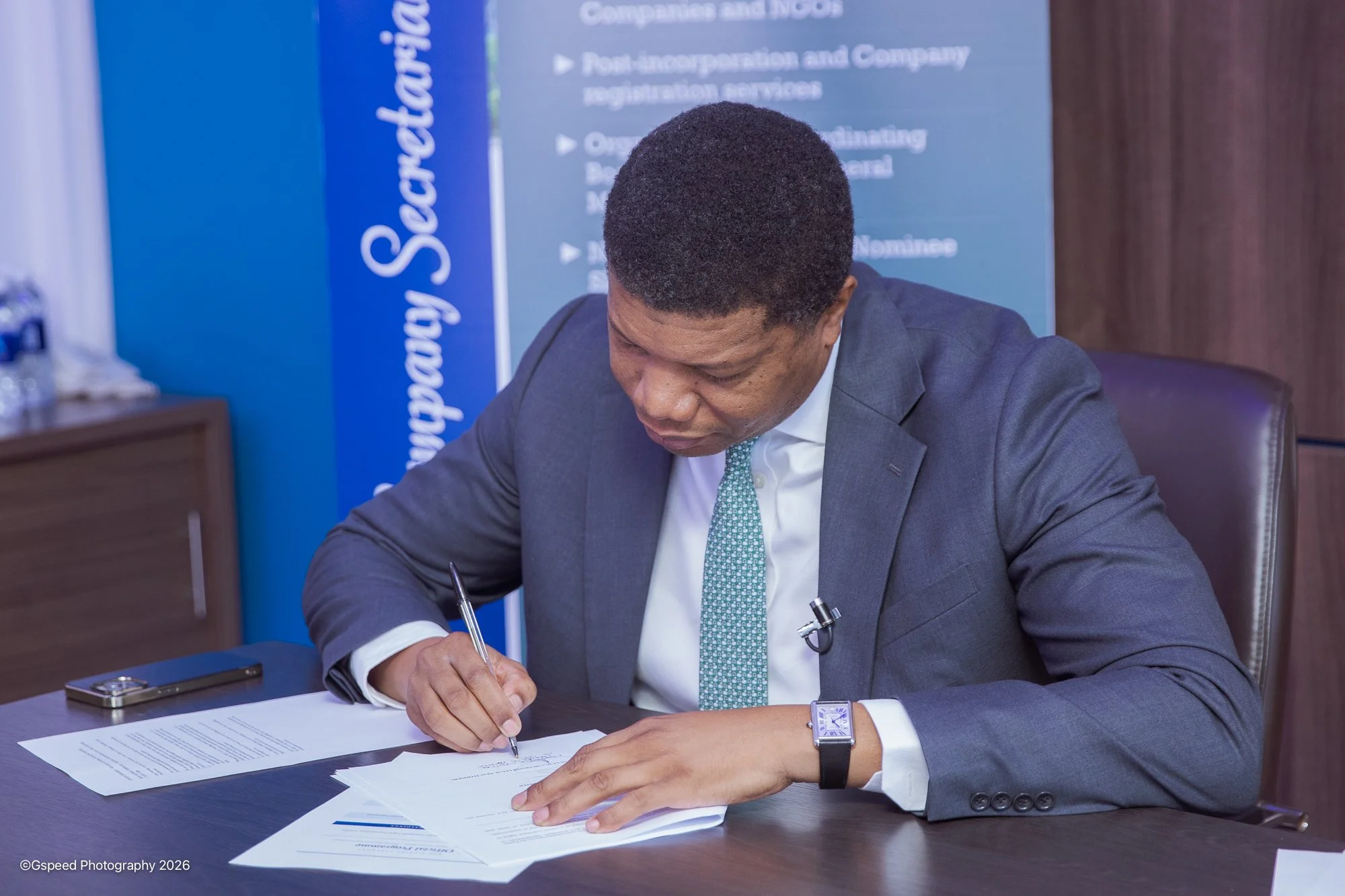 A man in a grey suit and white shirt seated at a desk, signing papers with a pen. He is wearing a watch on his wrist and a microphone clipped to his shirt. There are documents and a phone on the desk. Behind him, there is a blue display banner with w