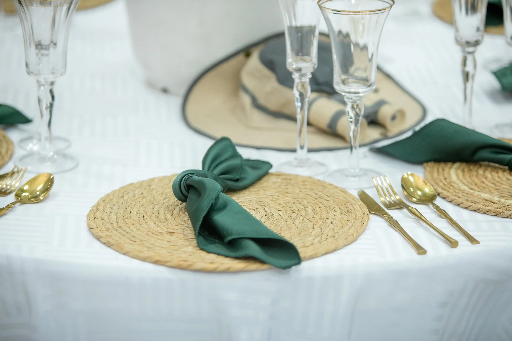 A table setting with woven placemats, green napkins tied with green fabric, gold utensils, clear glasses, and a beige and gray table runner.