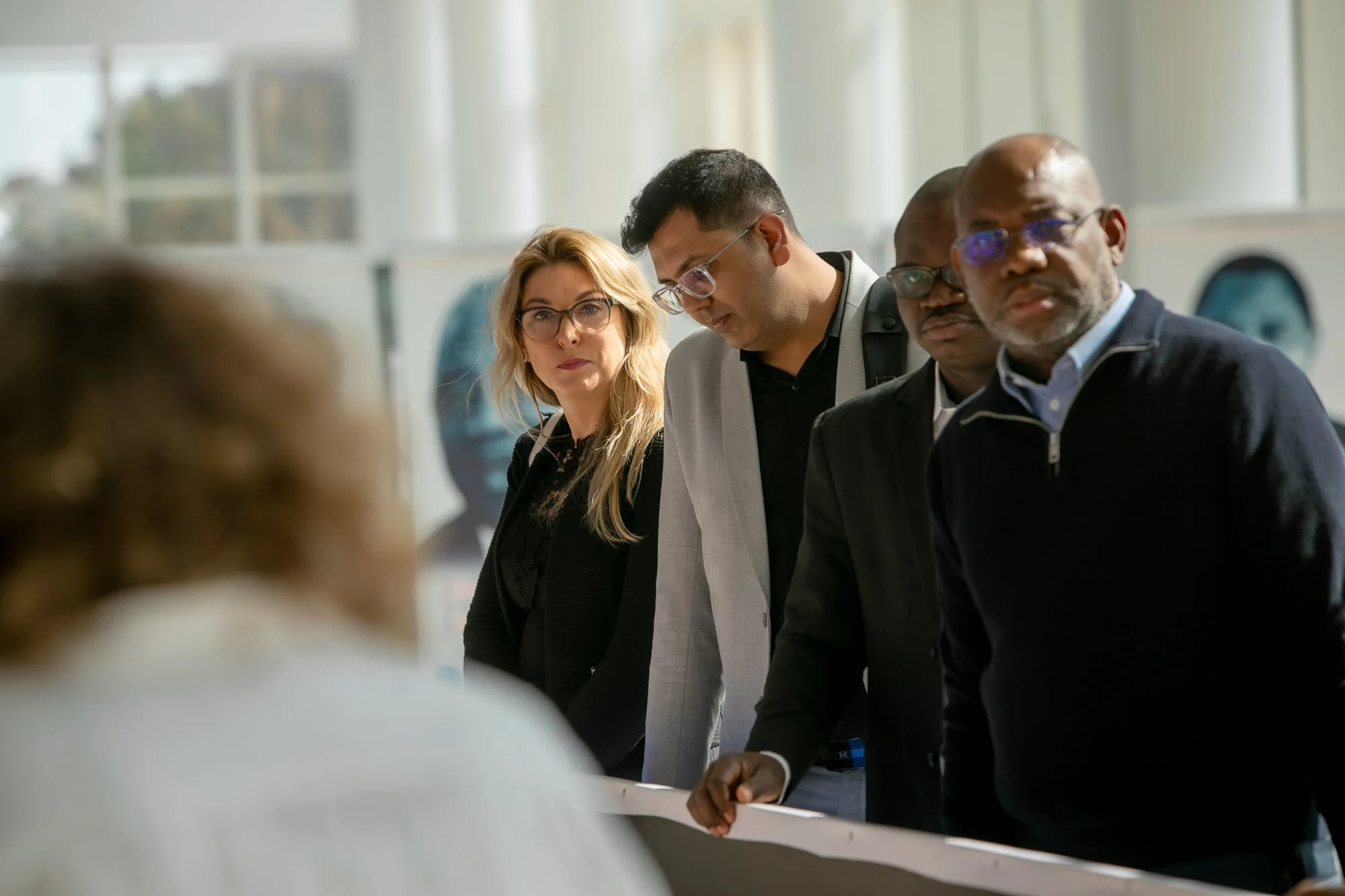 Group of diverse individuals in a meeting or discussion, leaning over a table, with a woman wearing glasses, and a man with gray glasses in focus.