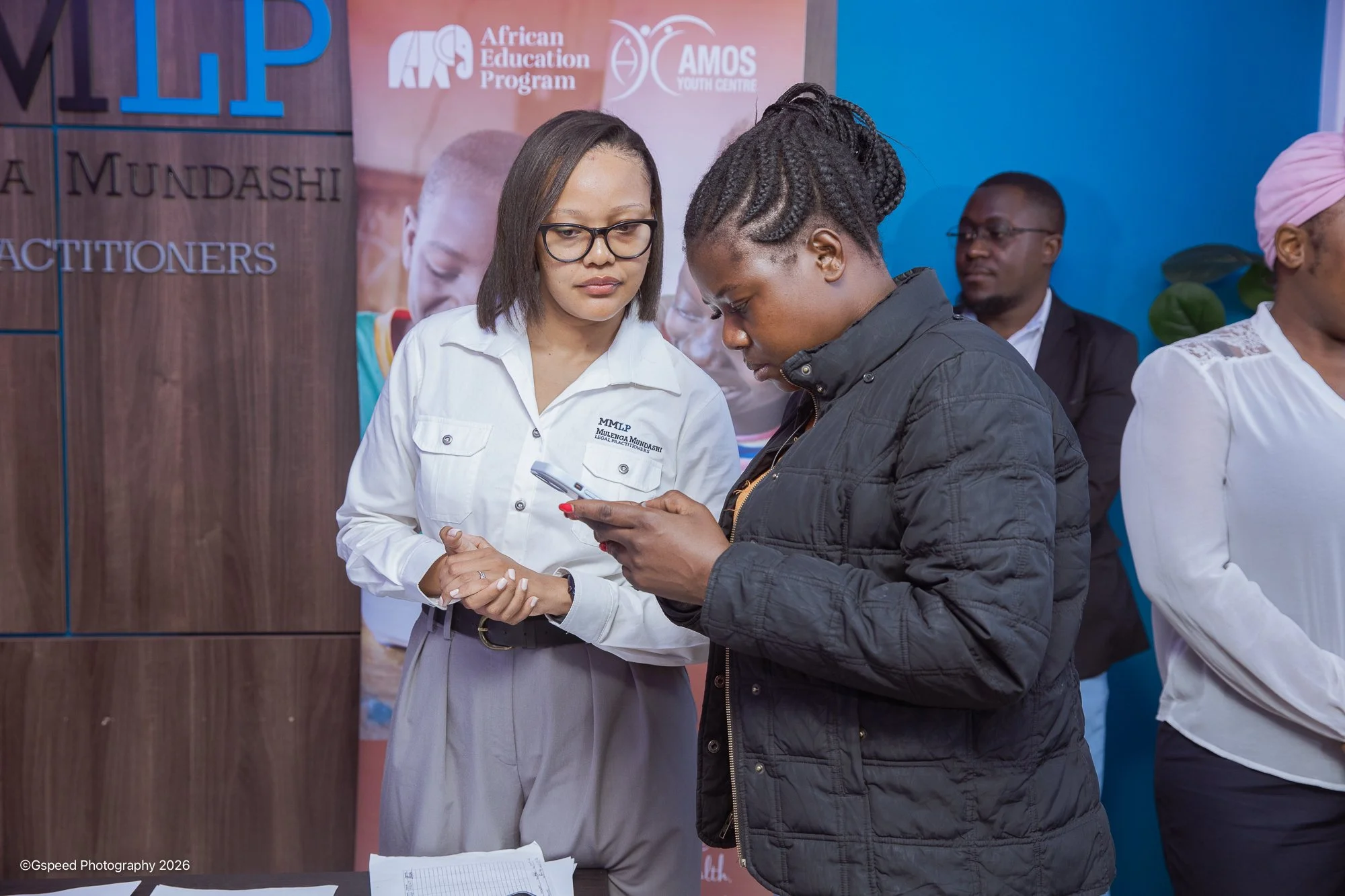 Two women looking at a phone together at an indoor event, with a man in the background and banners for the African Education Program and AMOS Youth Centre.