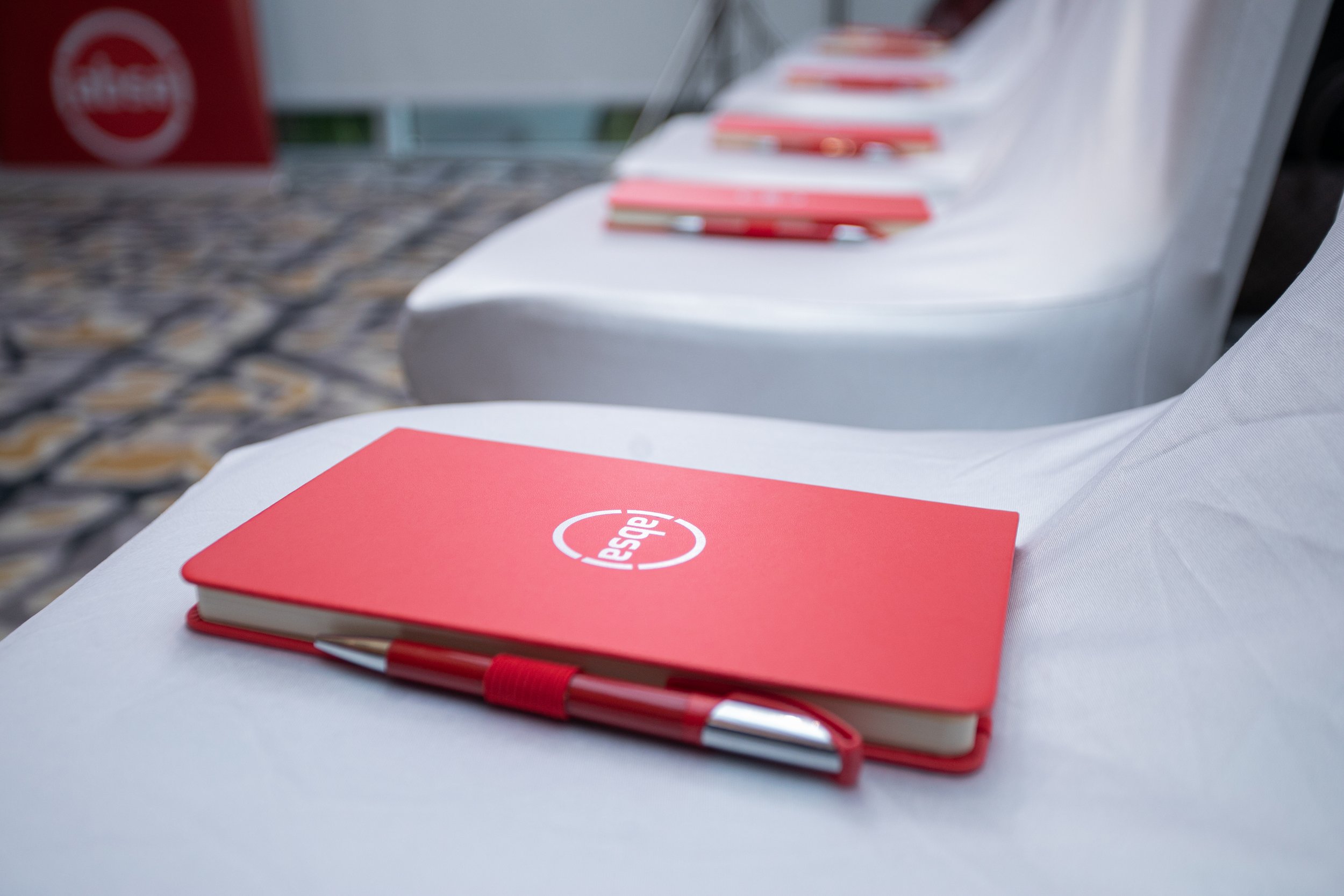 A red notebook with a white logo that says 'despegar' resting on a white chair. A red pen is clipped to the notebook. Other similar notebooks are visible in the background on the same chair.