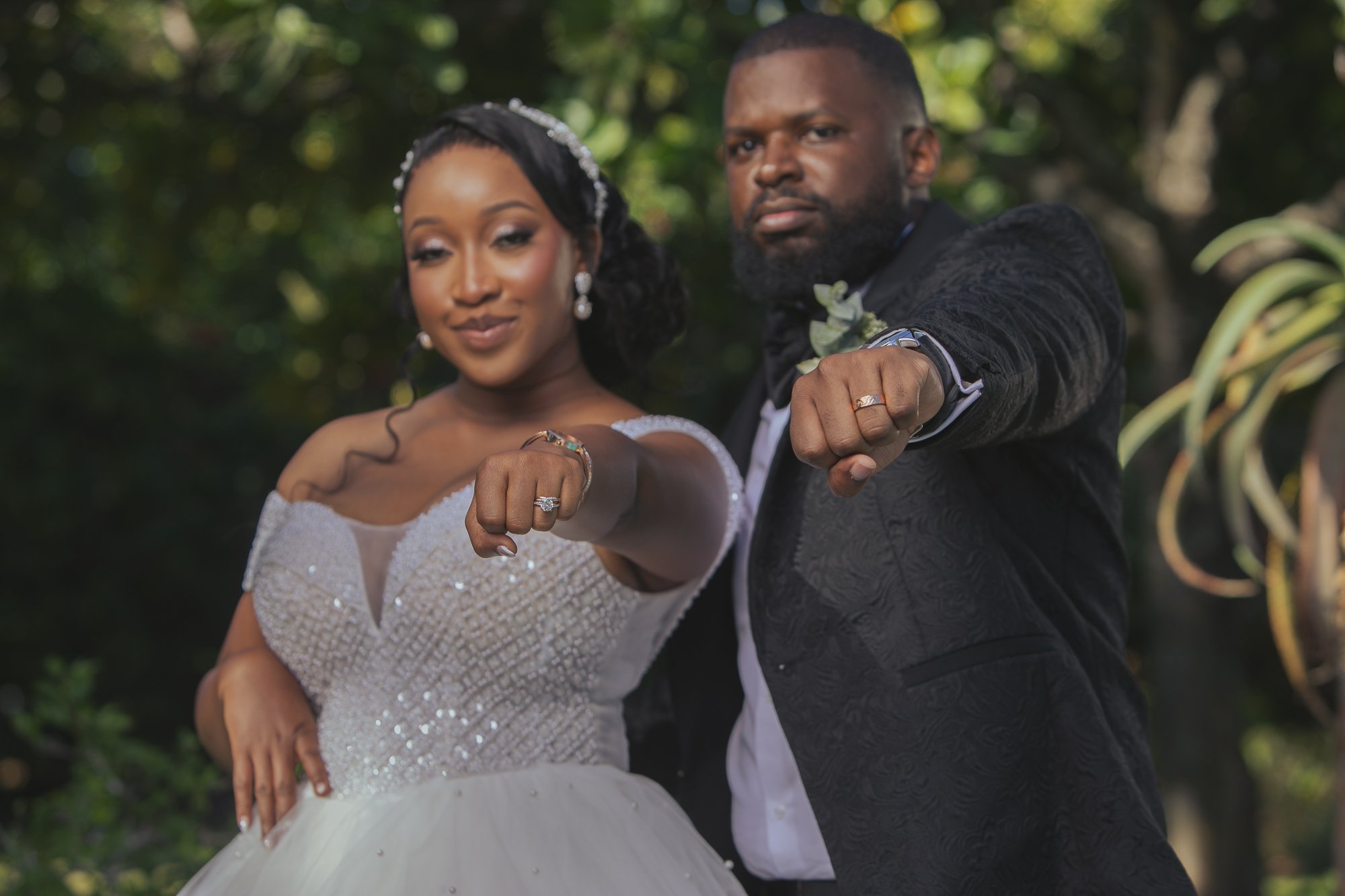 A bride and groom are outside in a lush green setting, showing their wedding rings and posing with their fists extended forward. The bride wears a white wedding gown with lace details, and the groom wears a black suit with a boutonniere. They both sm