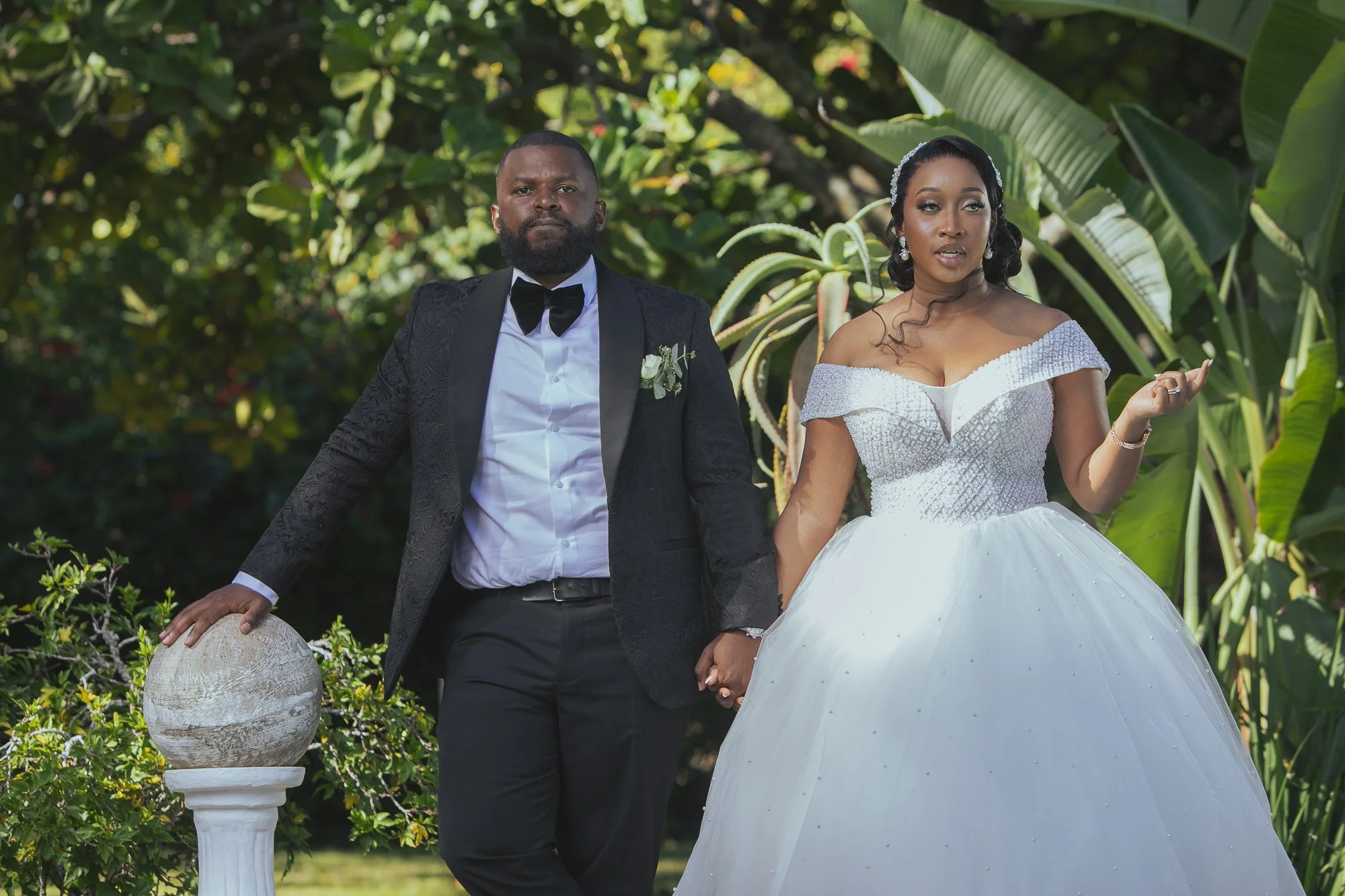A couple dressed in formal wedding attire standing outdoors among green foliage. The man is wearing a black tuxedo with a bow tie, and the woman is in a white wedding gown with off-the-shoulder sleeves and a tiara. They are holding hands, and the wom