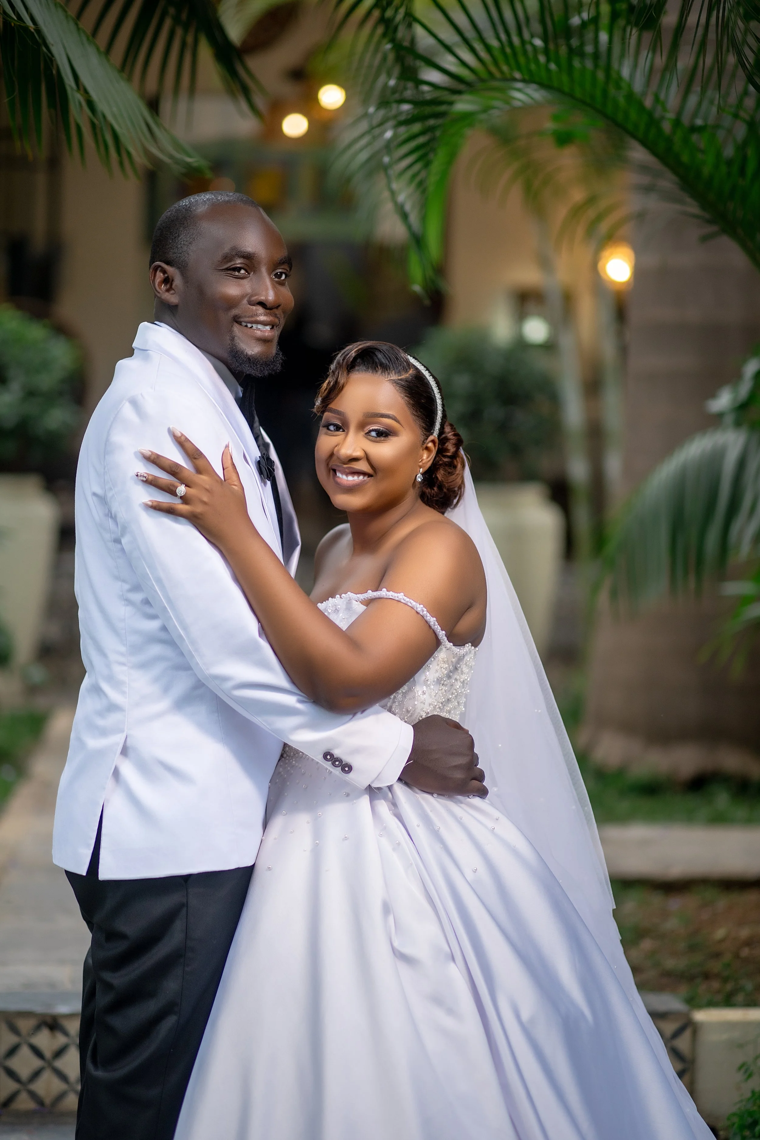 A newlywed couple embracing outdoors, surrounded by green palm leaves and blurred background lights, smiling warmly at the camera.