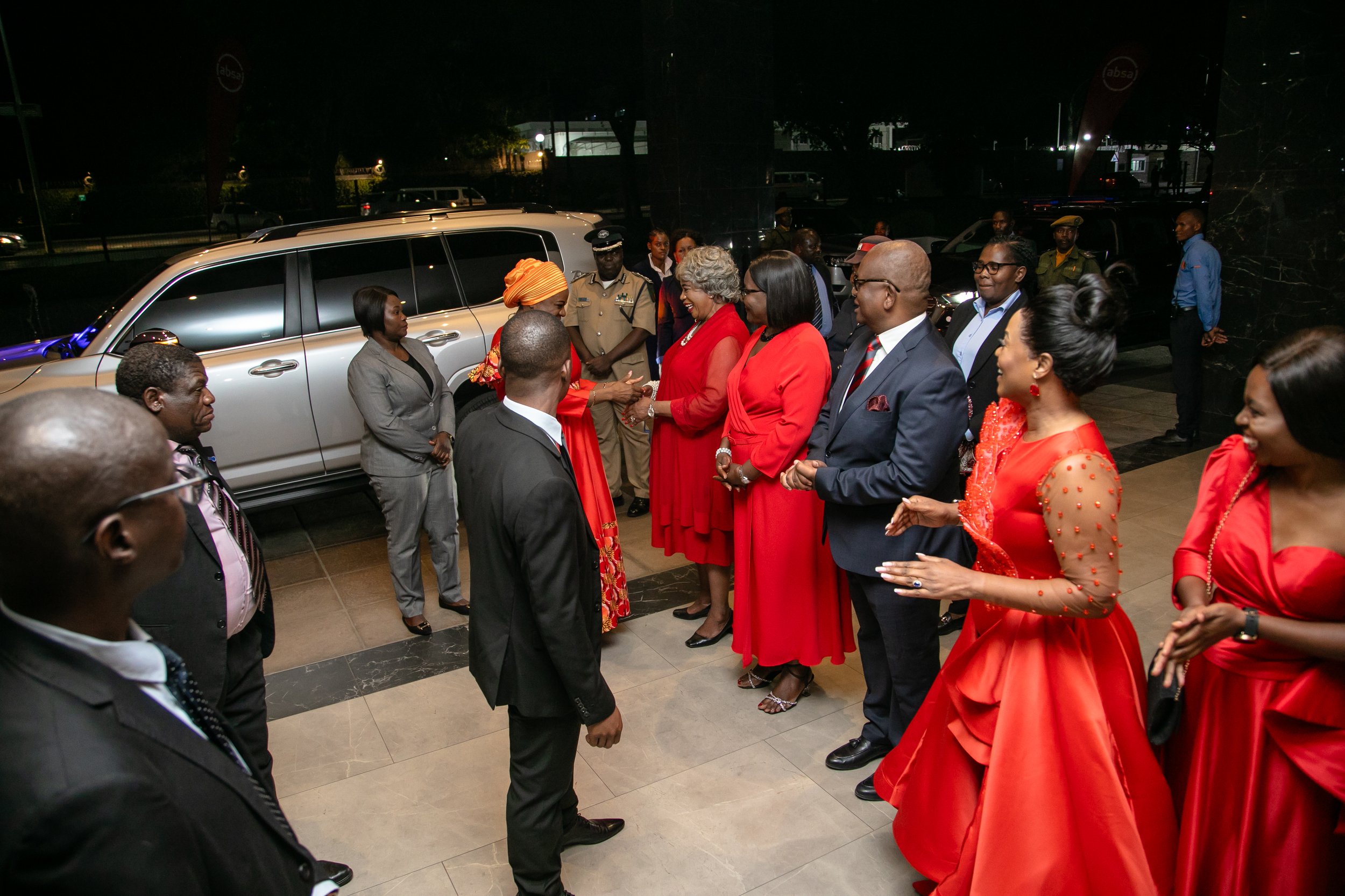 Group of people in formal attire gathered outside at night, engaged in conversation, with vehicles and dark buildings in the background.