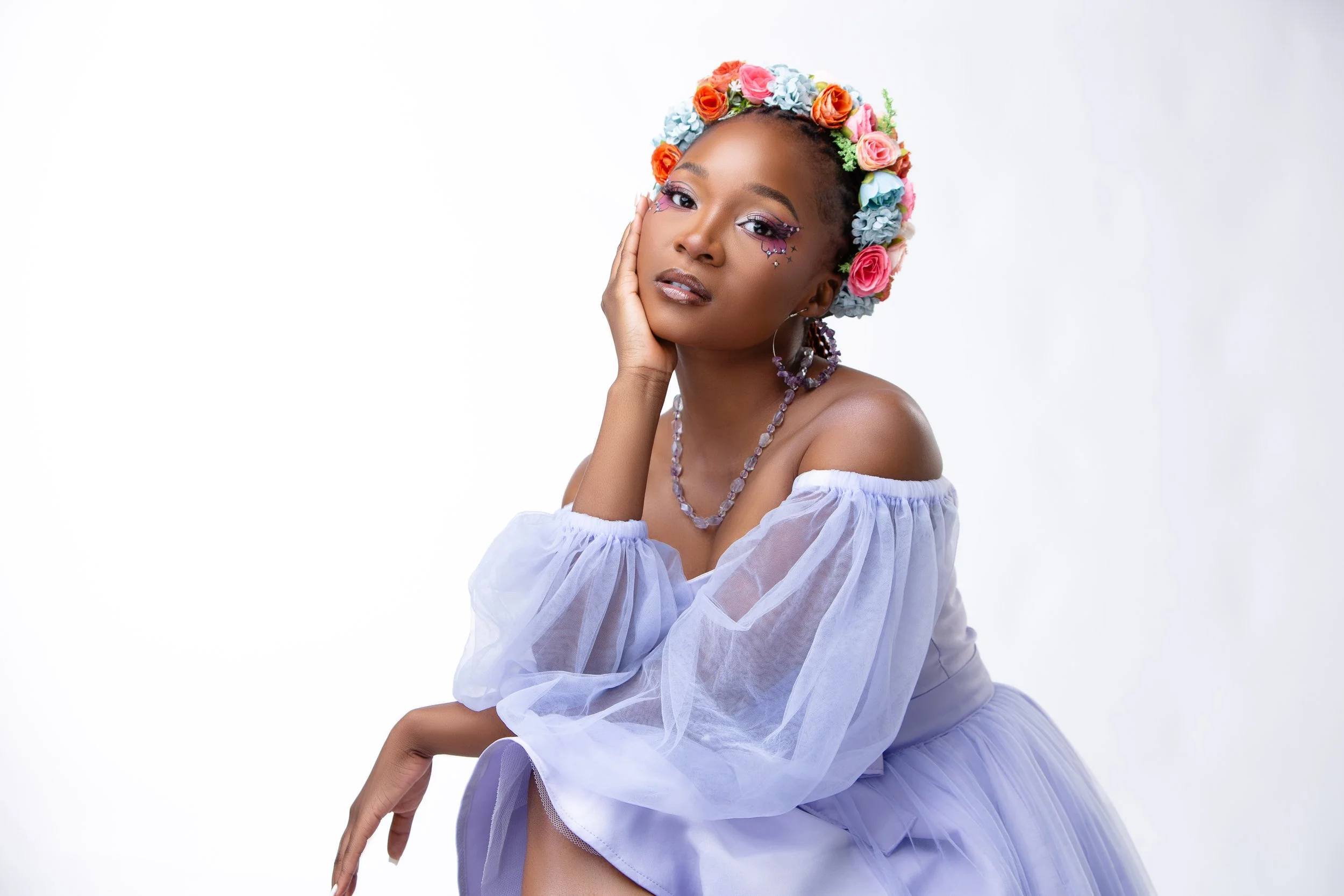 A woman with dark skin and braided hair wearing a floral crown of colorful roses and hydrangeas, with glitter makeup featuring stars, and purple jewelry, posing with her hand resting on her cheek against a white background.