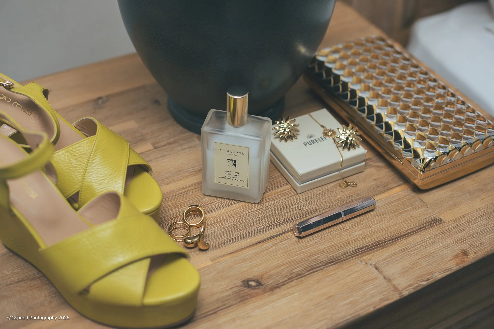 Yellow high-heeled shoes, gold jewelry including rings and earrings, a perfume bottle, a necklace, and a jewelry box with a geometric metallic design on a wooden surface.