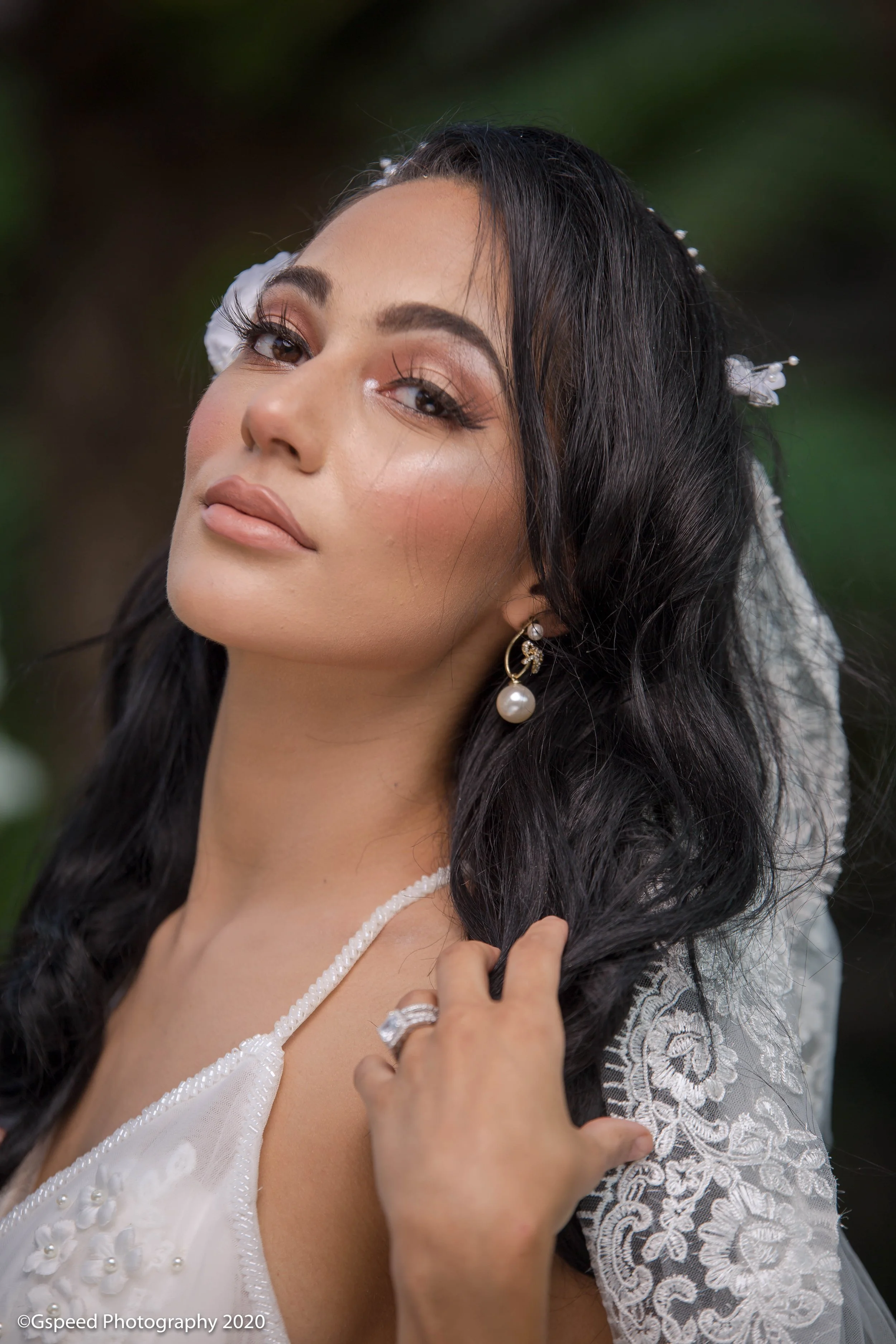 Close-up of a woman with dark hair, wearing pearl earrings and a white dress with floral embroidery, touching her hair with her hand.
