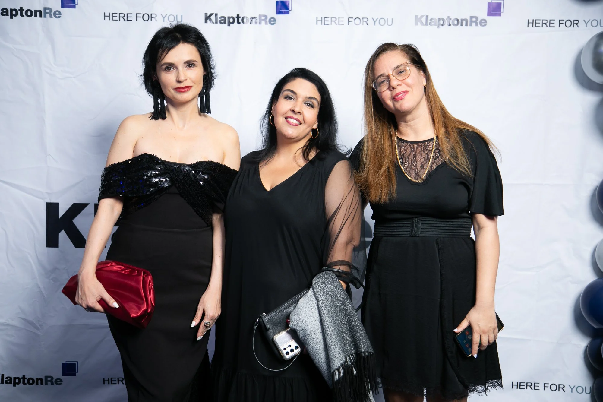 Three women in black dresses are posing together at an event with a backdrop that says 'KlaptonRe' and 'HERE FOR YOU.' They are smiling and holding bags and accessories.