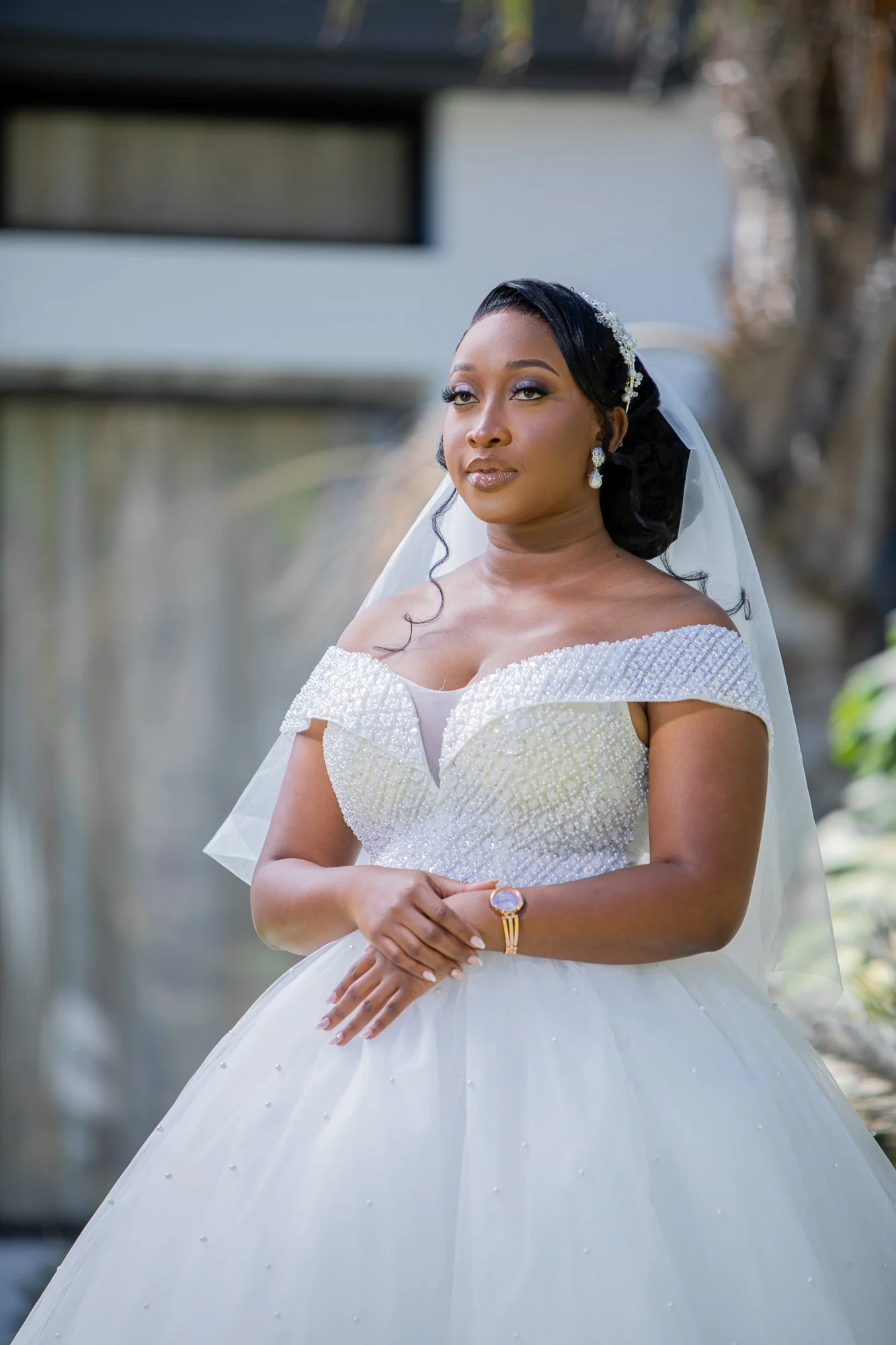 A bride standing outdoors in a wedding dress, with her arms crossed, wearing earrings and a bracelet, in a natural setting with blurred background.