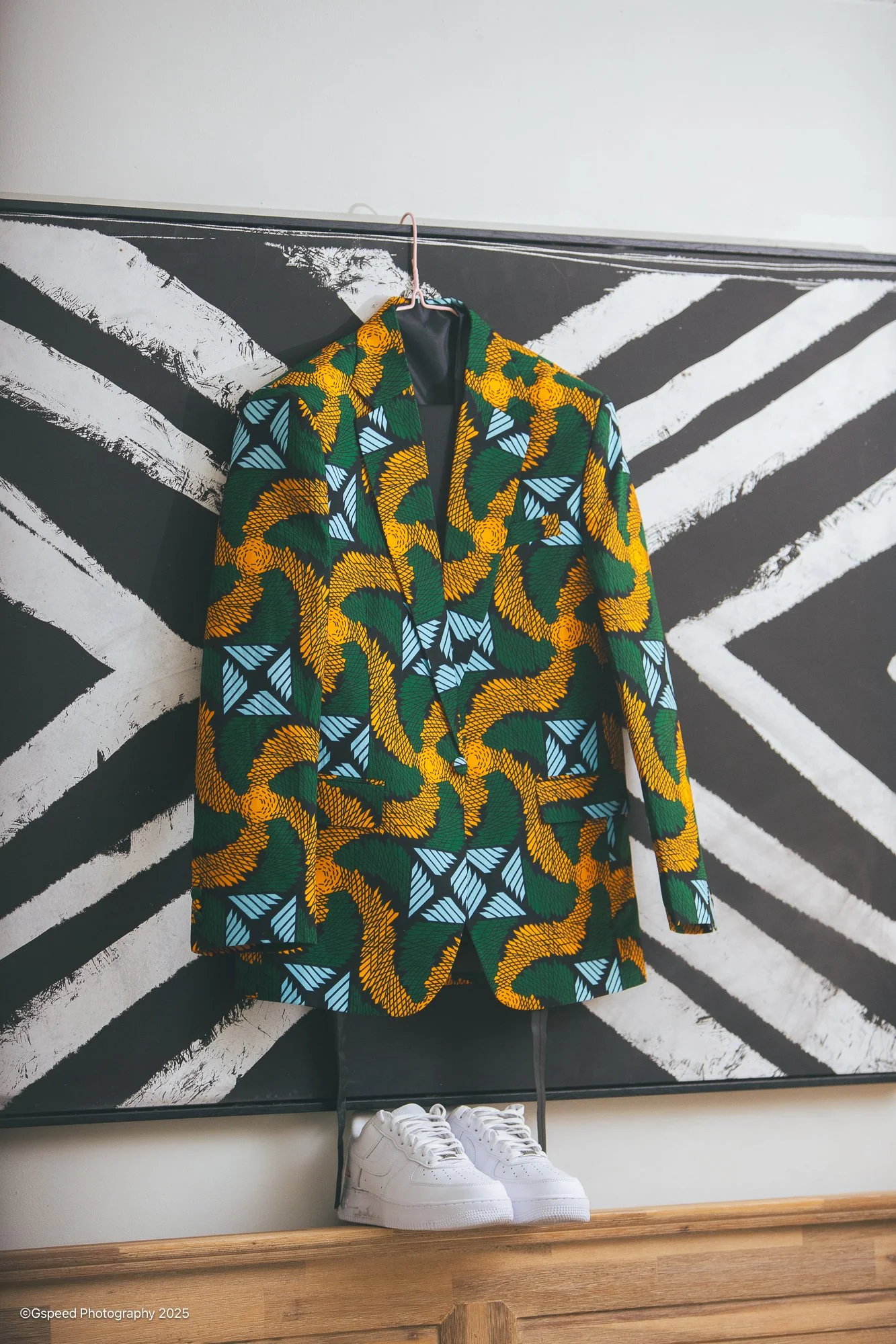 A colorful jacket with a bold pattern hangs on a hanger, with a pair of white sneakers placed below it. The backdrop features black and white painted diagonal stripes.