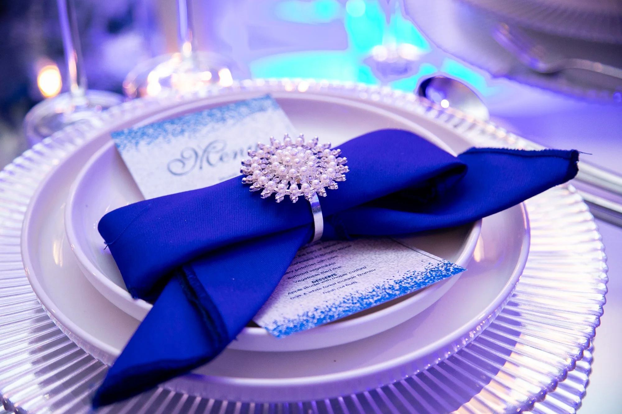Place setting with a dark blue napkin, pearl and rhinestone ring, silverware, menu, and a small snowflake-shaped ornament on a white plate, illuminated with blue and purple lighting.
