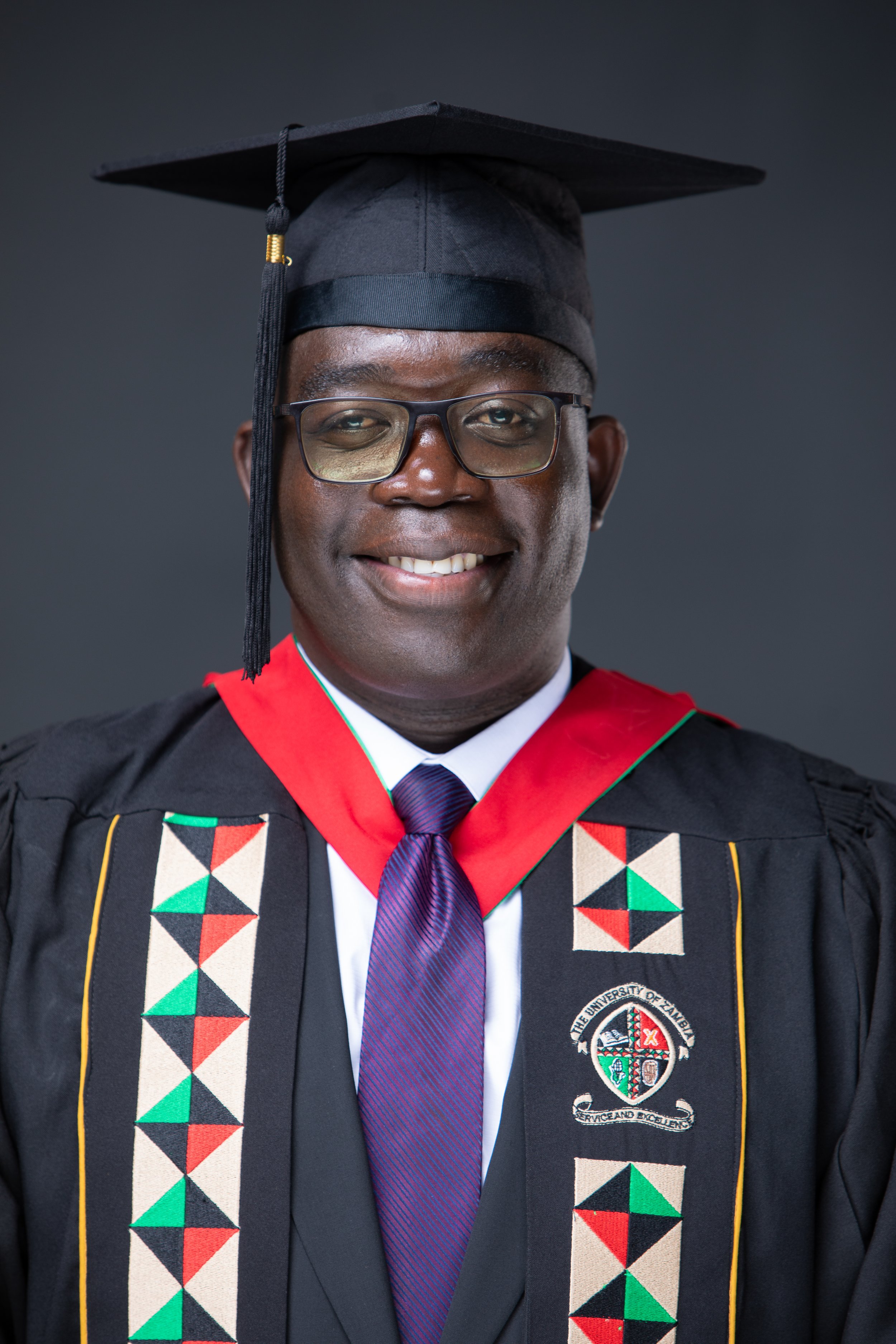 Graduation portrait of a man wearing academic regalia, including black gown, colorful patterned trim, and a black mortarboard cap, smiling in front of a dark background.