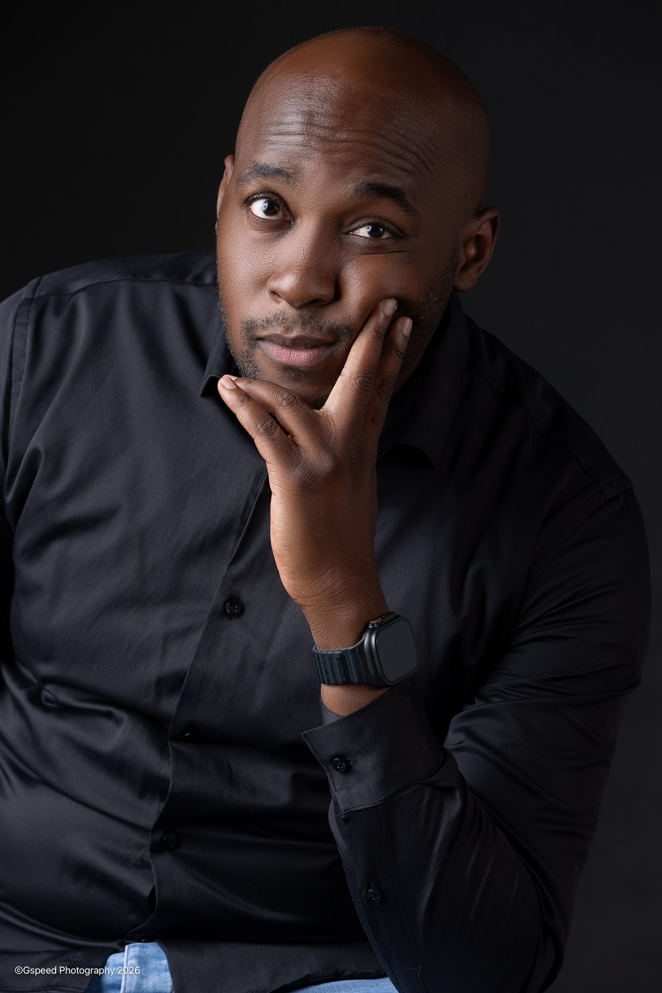 A portrait of a man with a shaved head wearing a black button-up shirt, looking at the camera with a thoughtful expression, resting his chin on his hand, against a black background.
