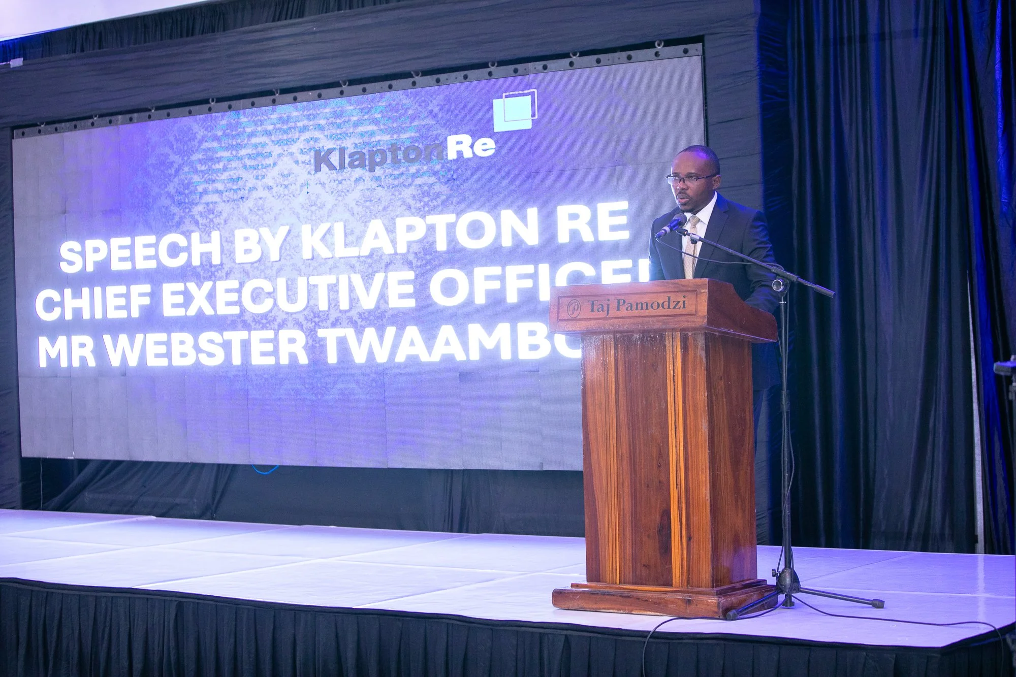 A man in a dark suit and glasses stands at a podium labeled 'Taj Pamodzi,' speaking at a conference or event. Behind him is a large screen displaying a presentation with the words 'Speech by Klapton Re, Chief Executive Officer Mr. Webster Twaambo' an