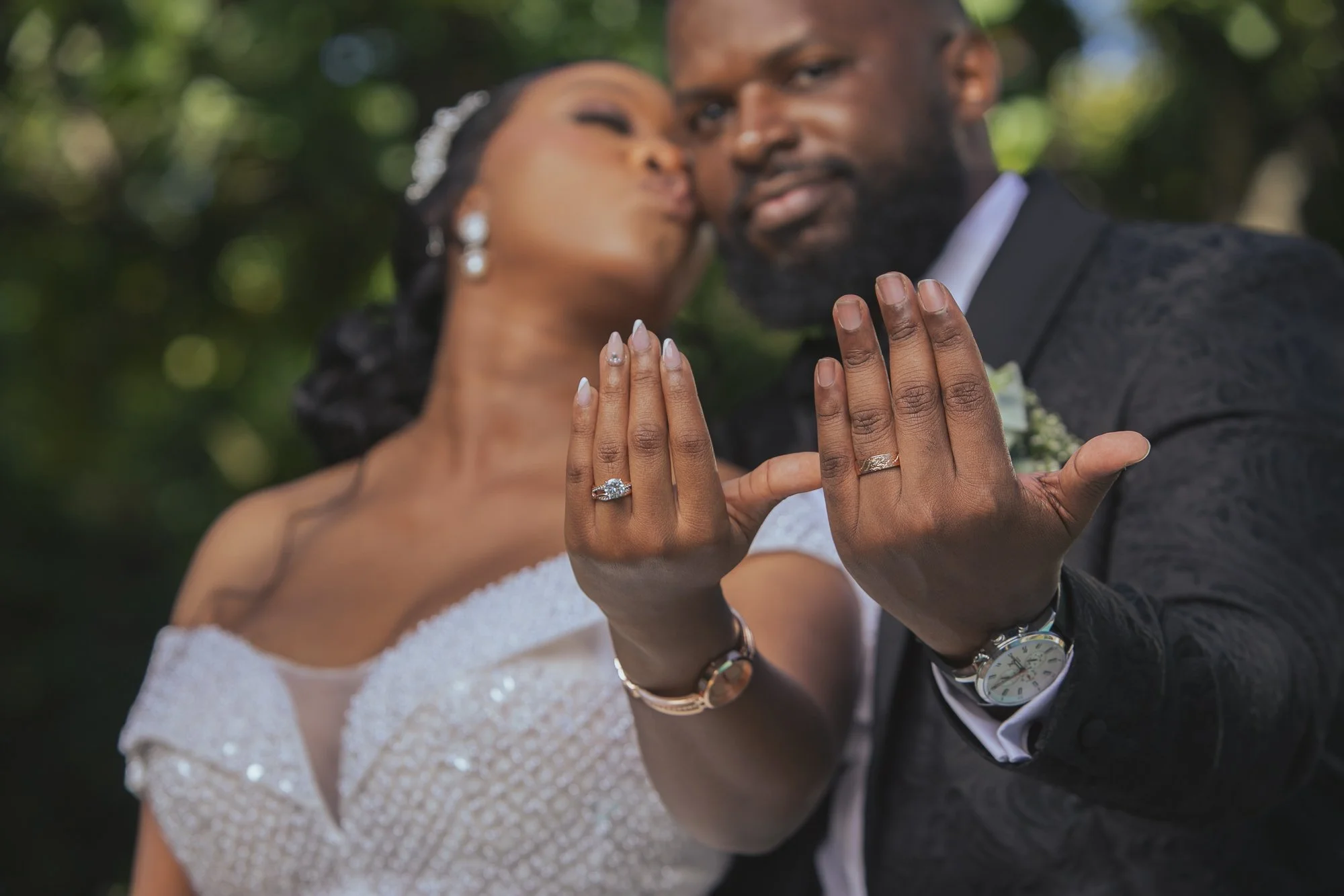 Close-up of a newly married couple displaying their wedding rings with a blurred outdoor background.