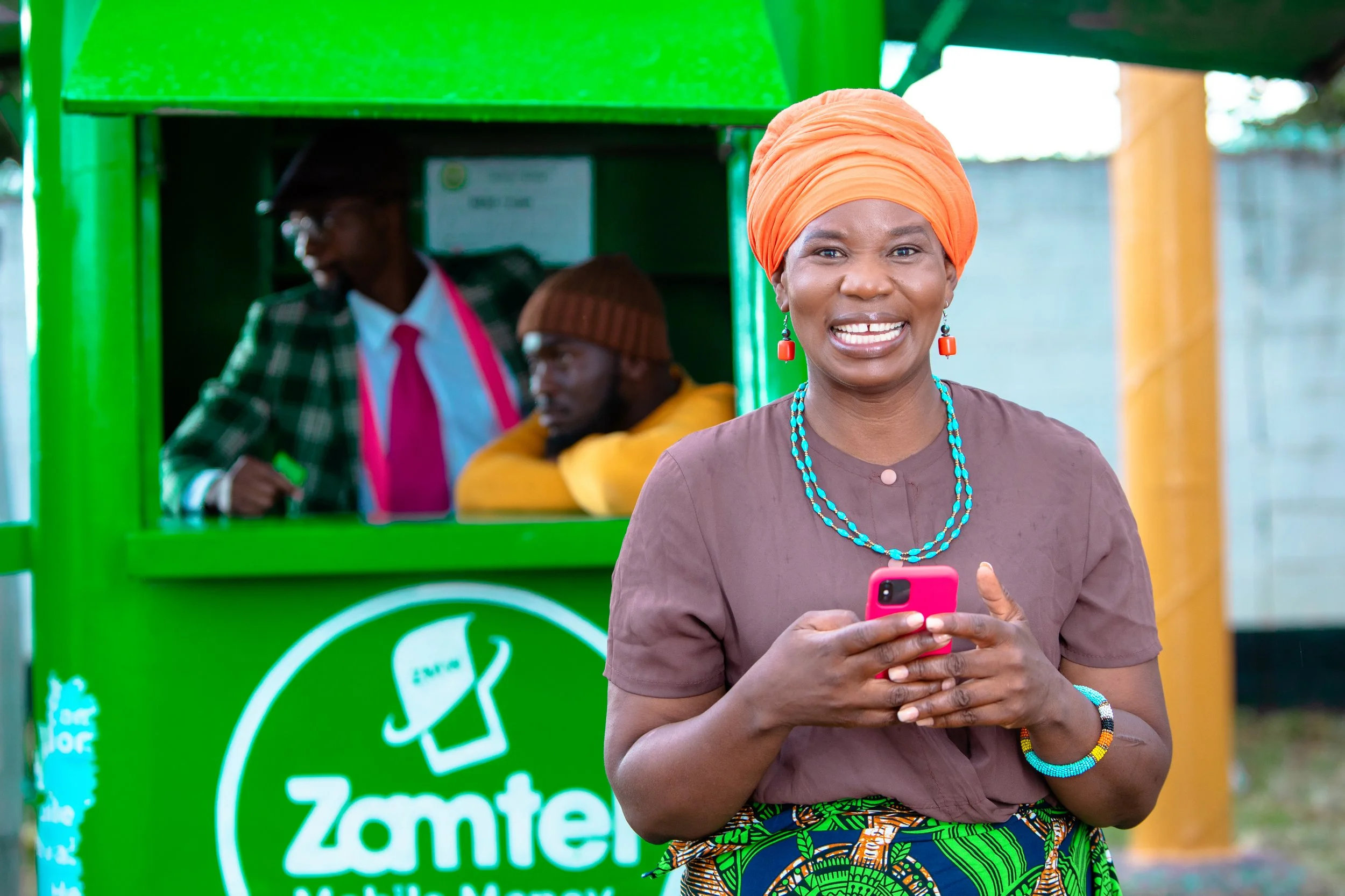 A woman wearing an orange headwrap and jewelry, smiling and holding a pink smartphone, standing in front of a green Zamtel kiosk with other people in the background.