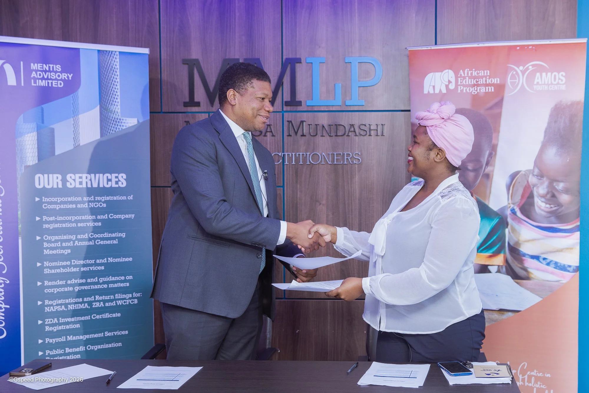 A man in a suit shaking hands with a woman in a white blouse and pink headwrap, both smiling, in front of a wall with 'MIMP' signage. There are banners and documents on the table.