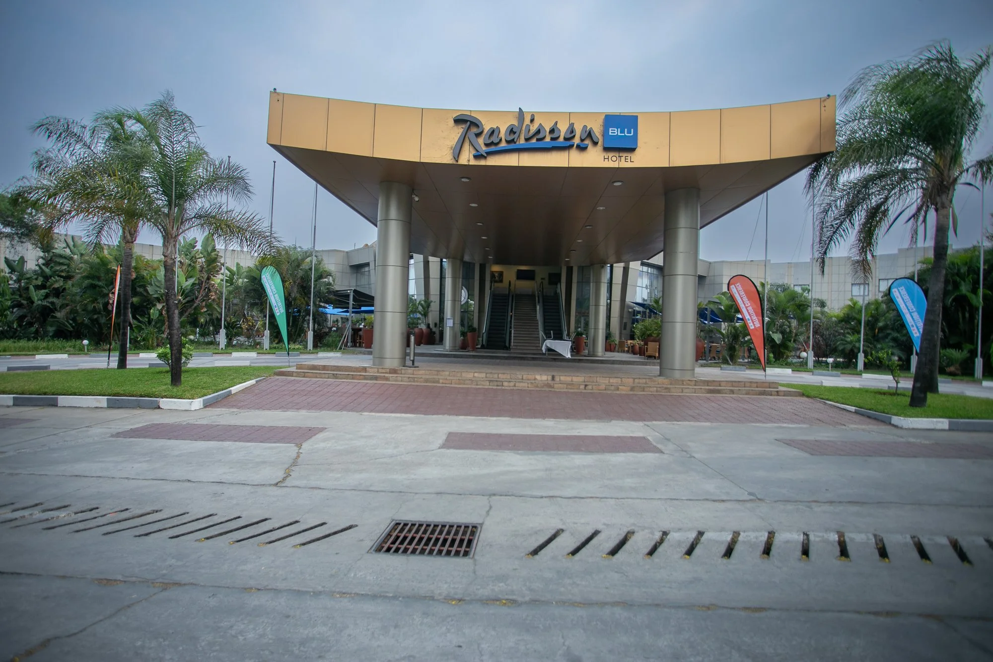 Entrance of a Radisson Blu hotel with a canopy, palm trees, and flags on either side, under a cloudy sky.