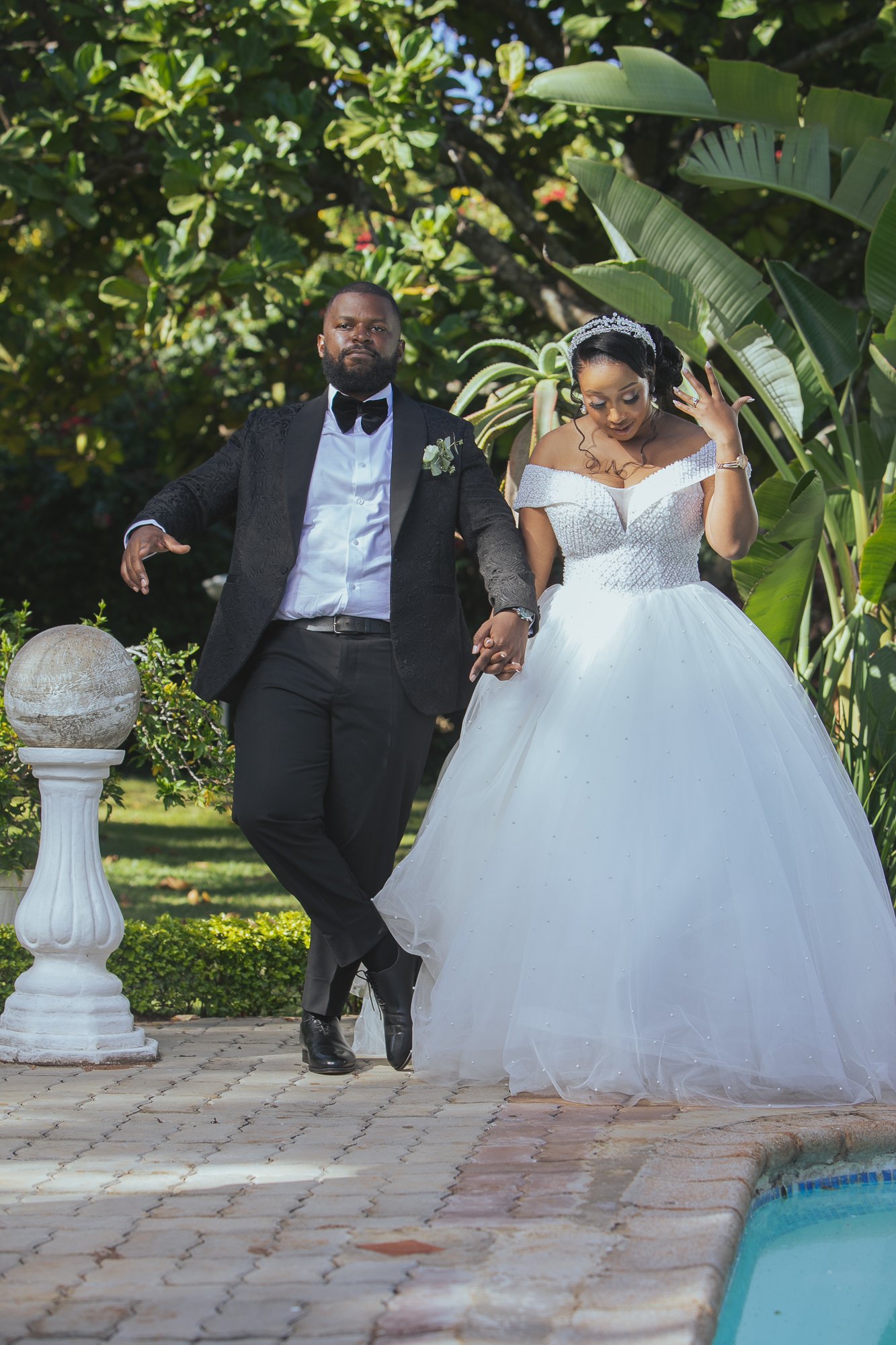 A bride and groom in wedding attire standing outdoors by a pool and lush greenery. The groom wears a tuxedo with a bow tie, and the bride wears a white wedding gown with off-the-shoulder sleeves and a tiara. They hold hands.