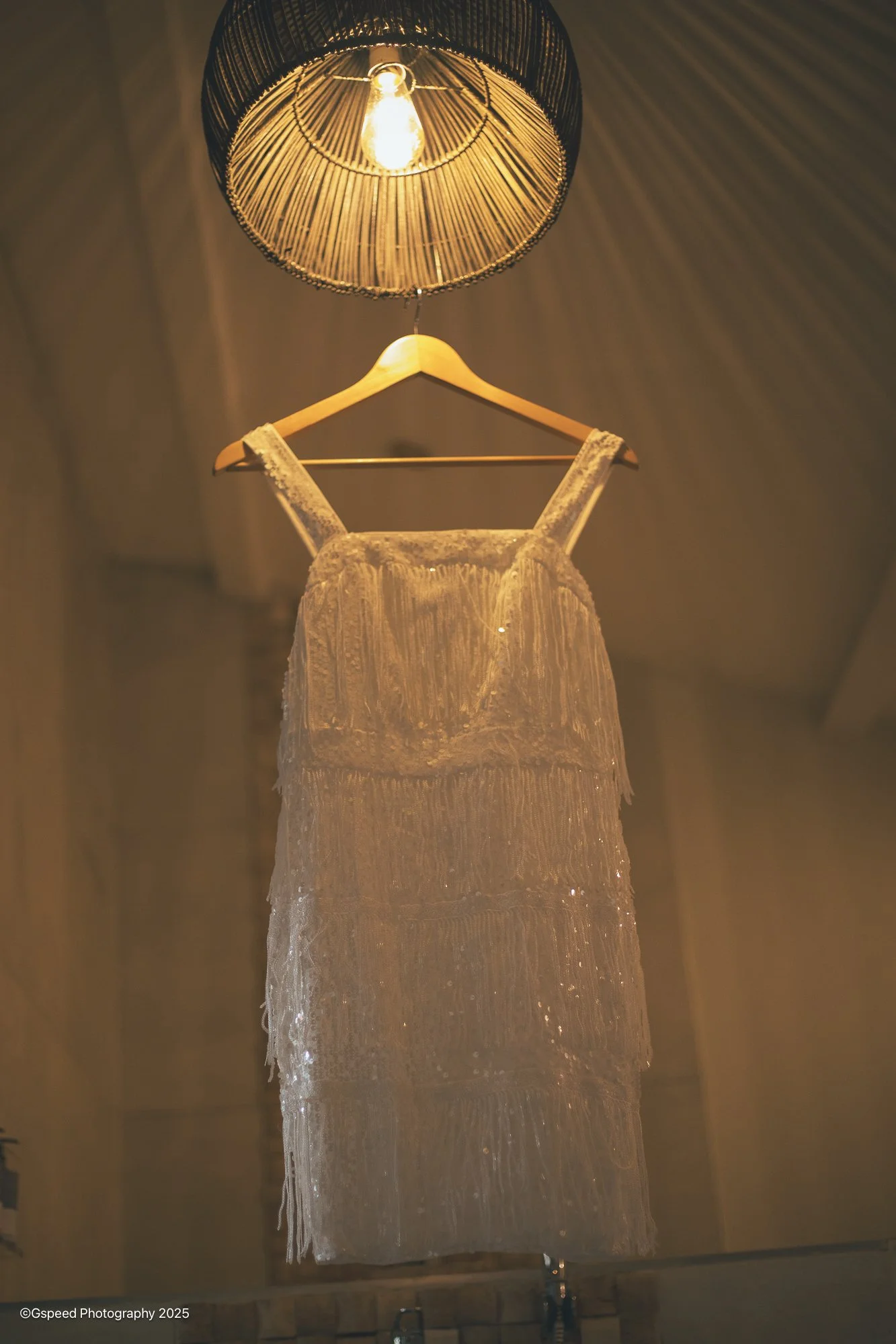 A white, sleeveless, lace dress hanging on a hanger under a lit light fixture with a lampshade made of wood or bamboo, in a softly lit room.