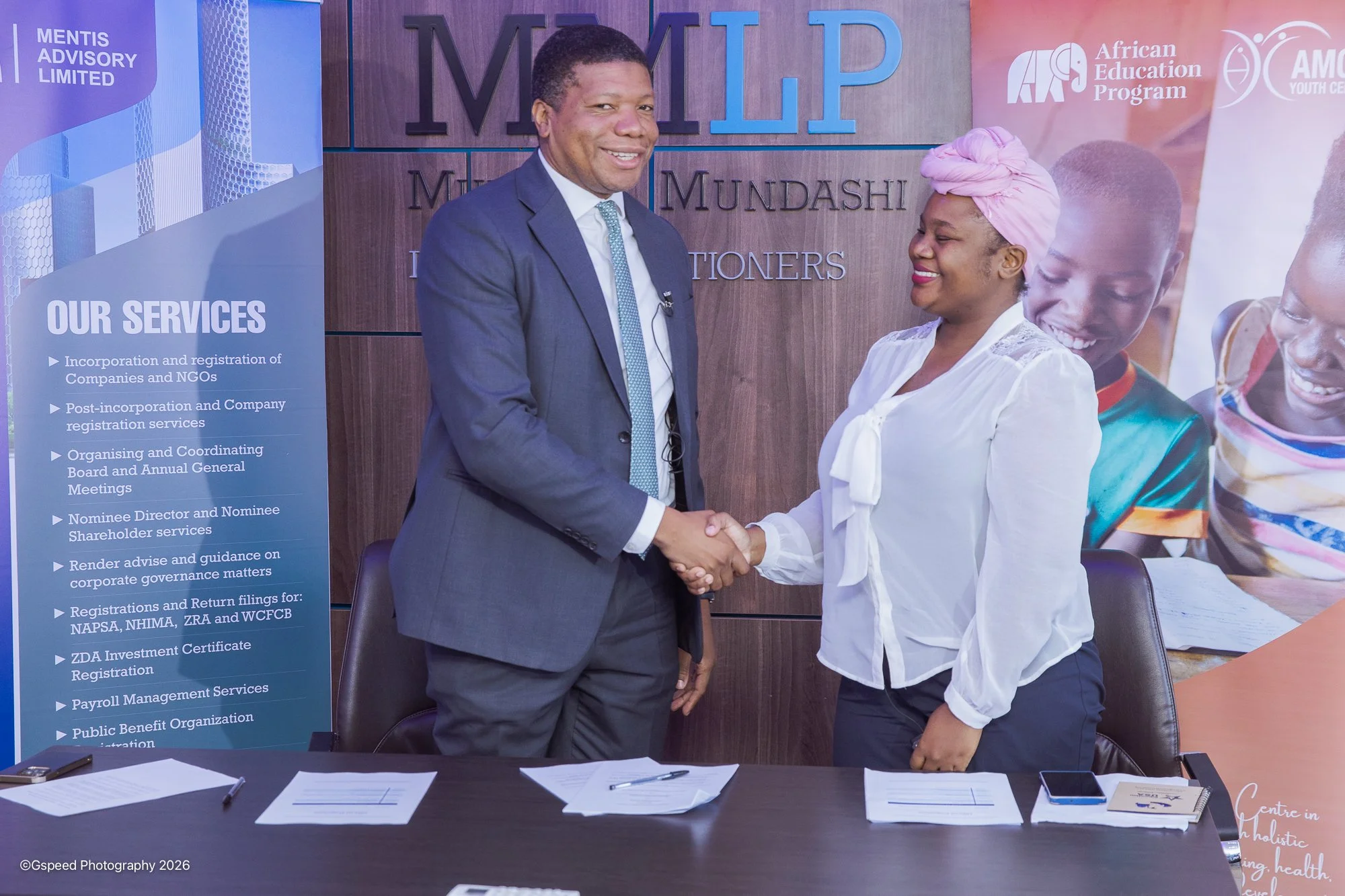 Two people, a man and a woman, shaking hands in a professional setting. The man is wearing a dark suit with a white shirt and dotted tie, and the woman is dressed in a white blouse with a pink headwrap. They are standing in front of a wooden wall wit