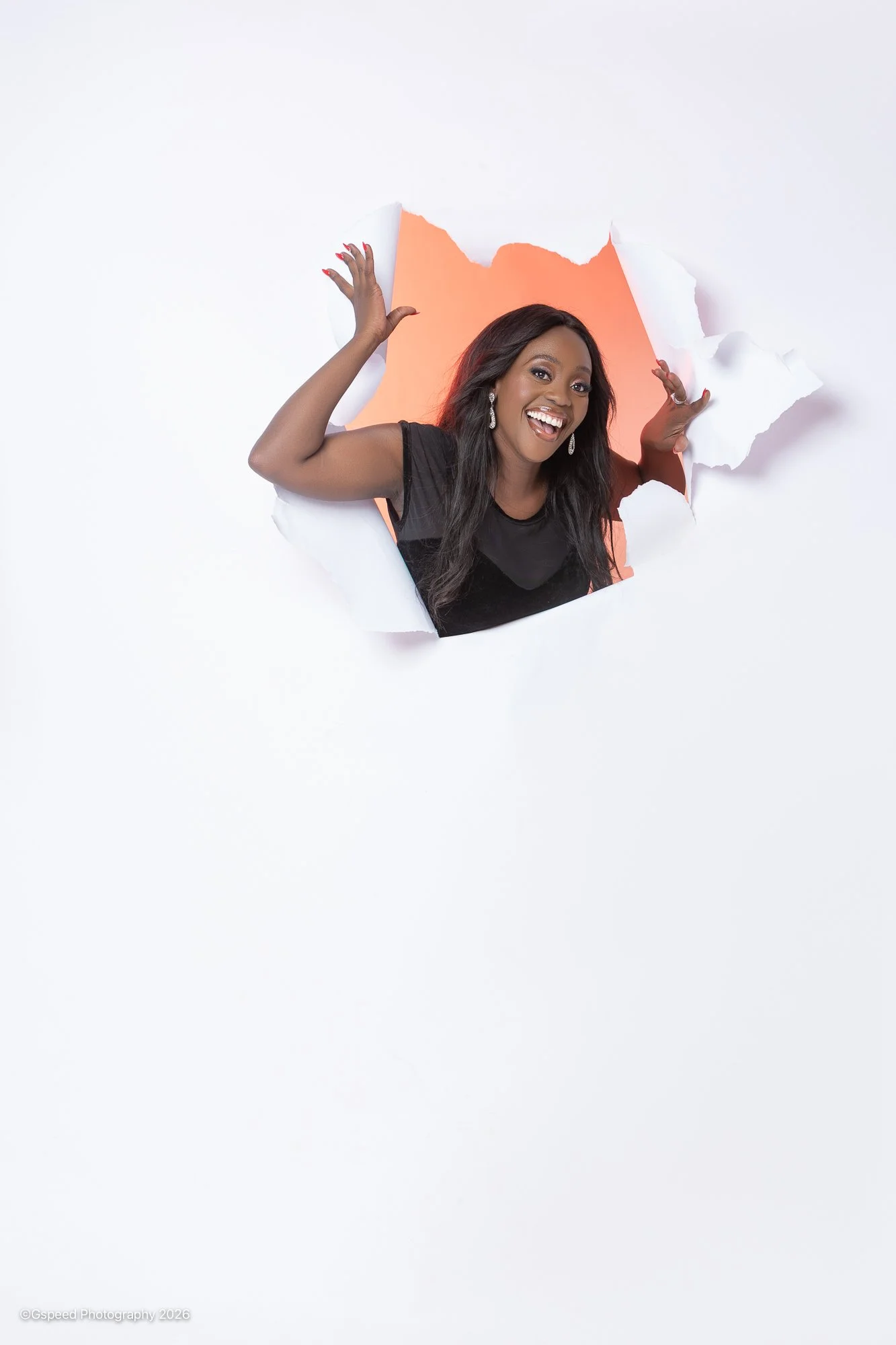 A woman with long dark hair and earrings smiling and poking her head and hands through a torn paper wall with a warm glowing backdrop.