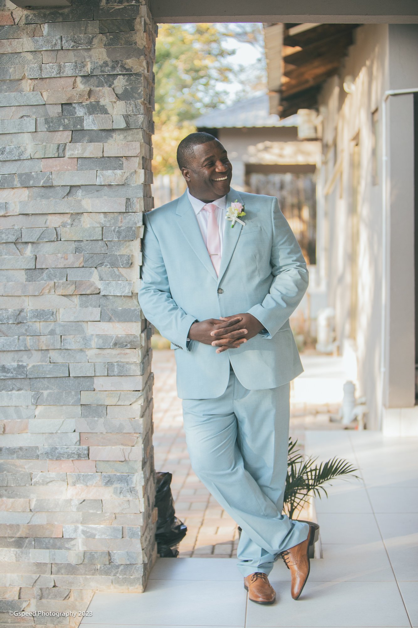 A man in a light blue suit, pink tie, and brown dress shoes stands outdoors, smiling and leaning against a stone wall. He is wearing a boutonniere with a pink rose and white flowers, with trees and a building in the background.