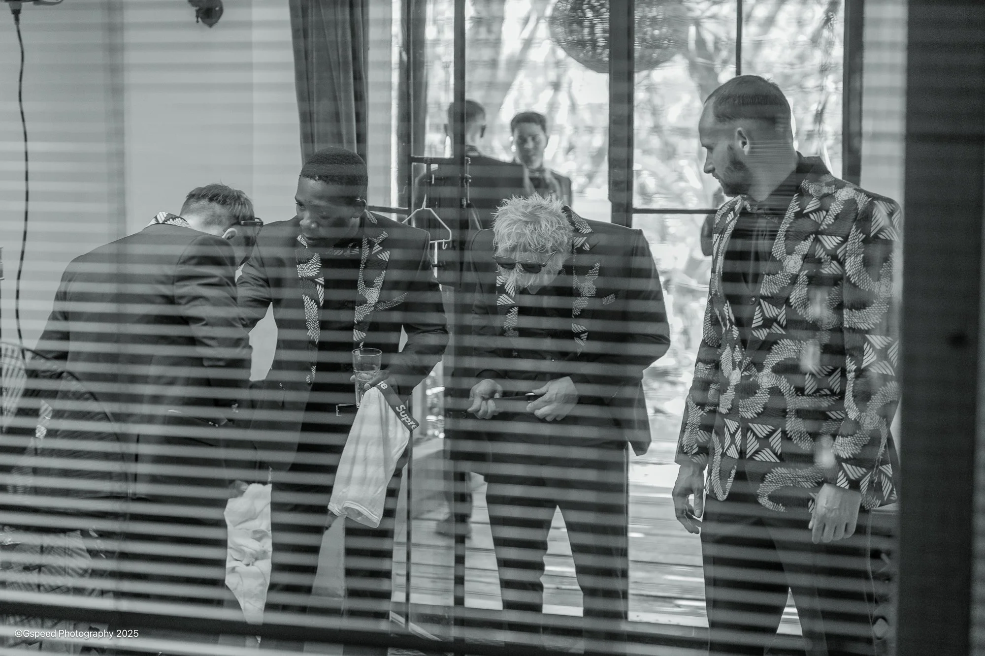 A group of five people wearing matching patterned suits, gathered around a table in a room with a large glass window. Some are examining items on the table, while others stand nearby.