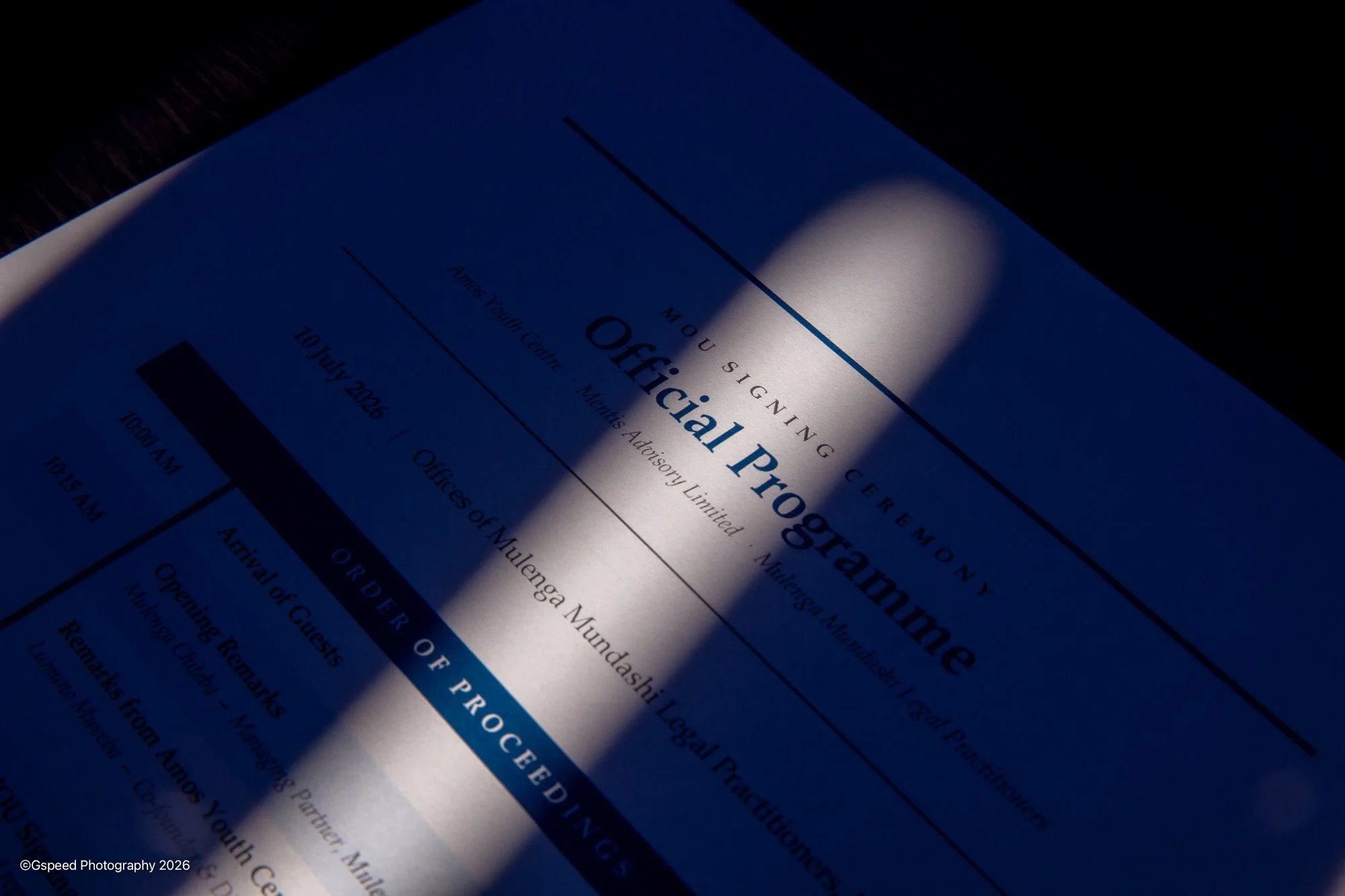 A printed document titled 'Official Program' with a date of July 19, 2026, partially illuminated by a focused light, with text about a conference or event on a dark surface.