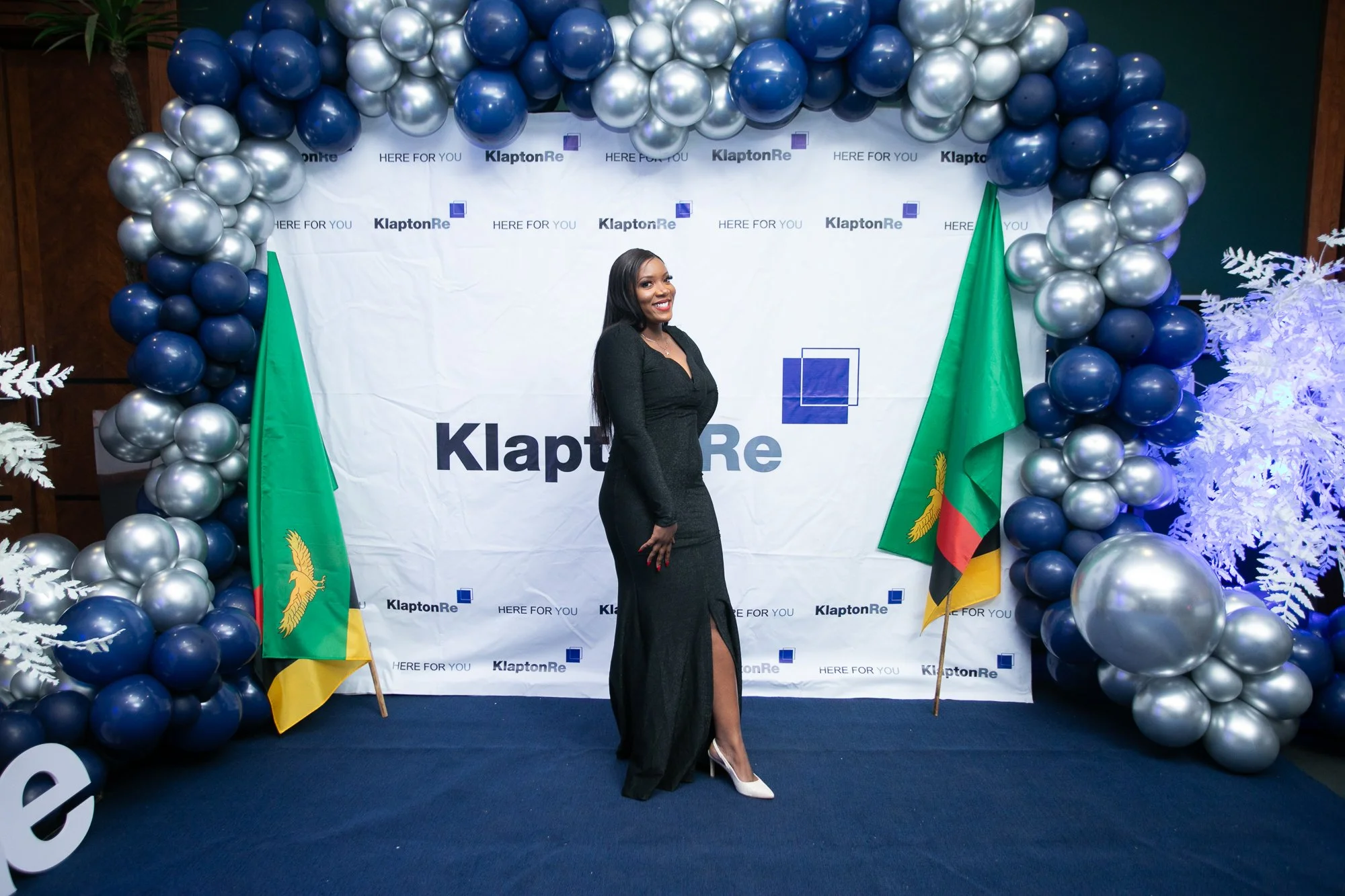 A woman in a black dress and white heels standing in front of a balloon arch and a backdrop with the KlaptonRe logo, national flags, and the phrase 'Here For You' at an event or celebration.