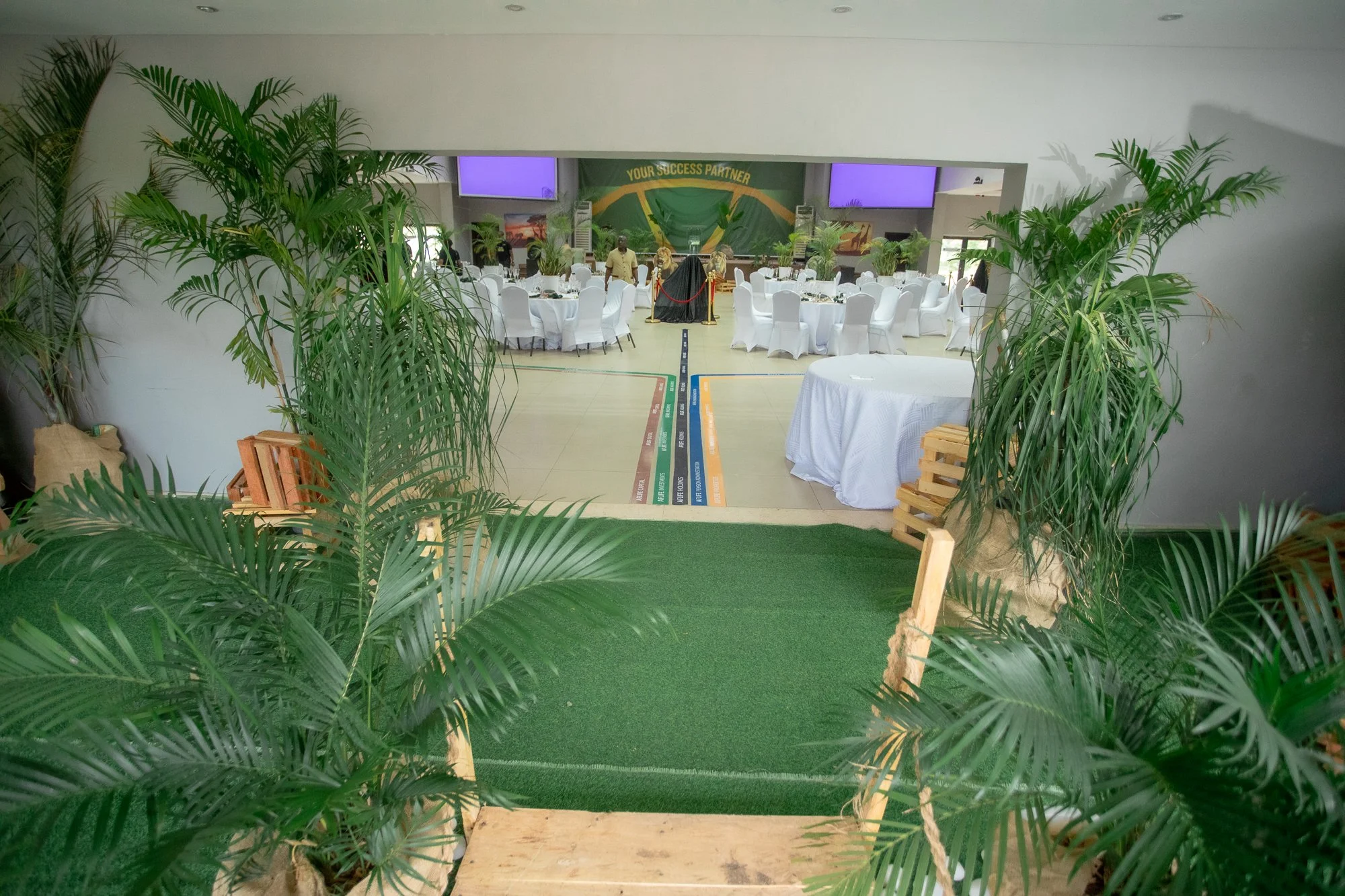 An event hall decorated with round tables covered with white tablecloths and white chairs, adorned with plant decorations. The hall has a stage area with a green backdrop and two purple screens. A line of colored markers or banners is on the floor le