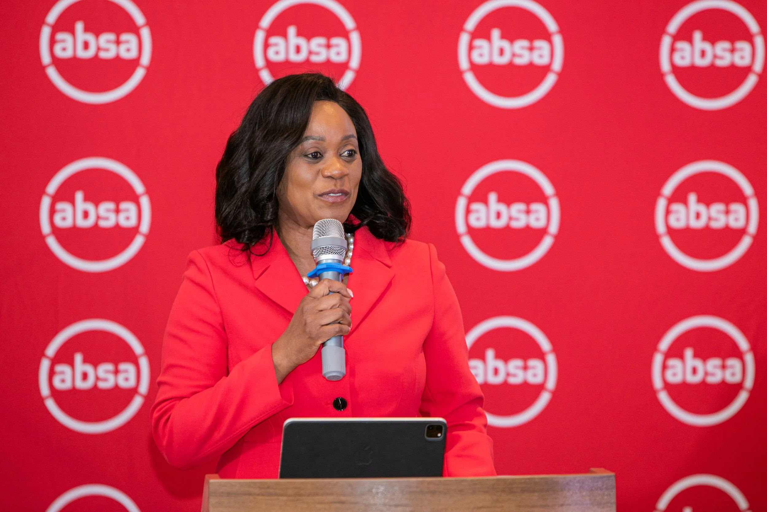 A woman in a red suit holding a microphone, standing at a podium against a red backdrop with the 'absa' logo repeating in white circles.