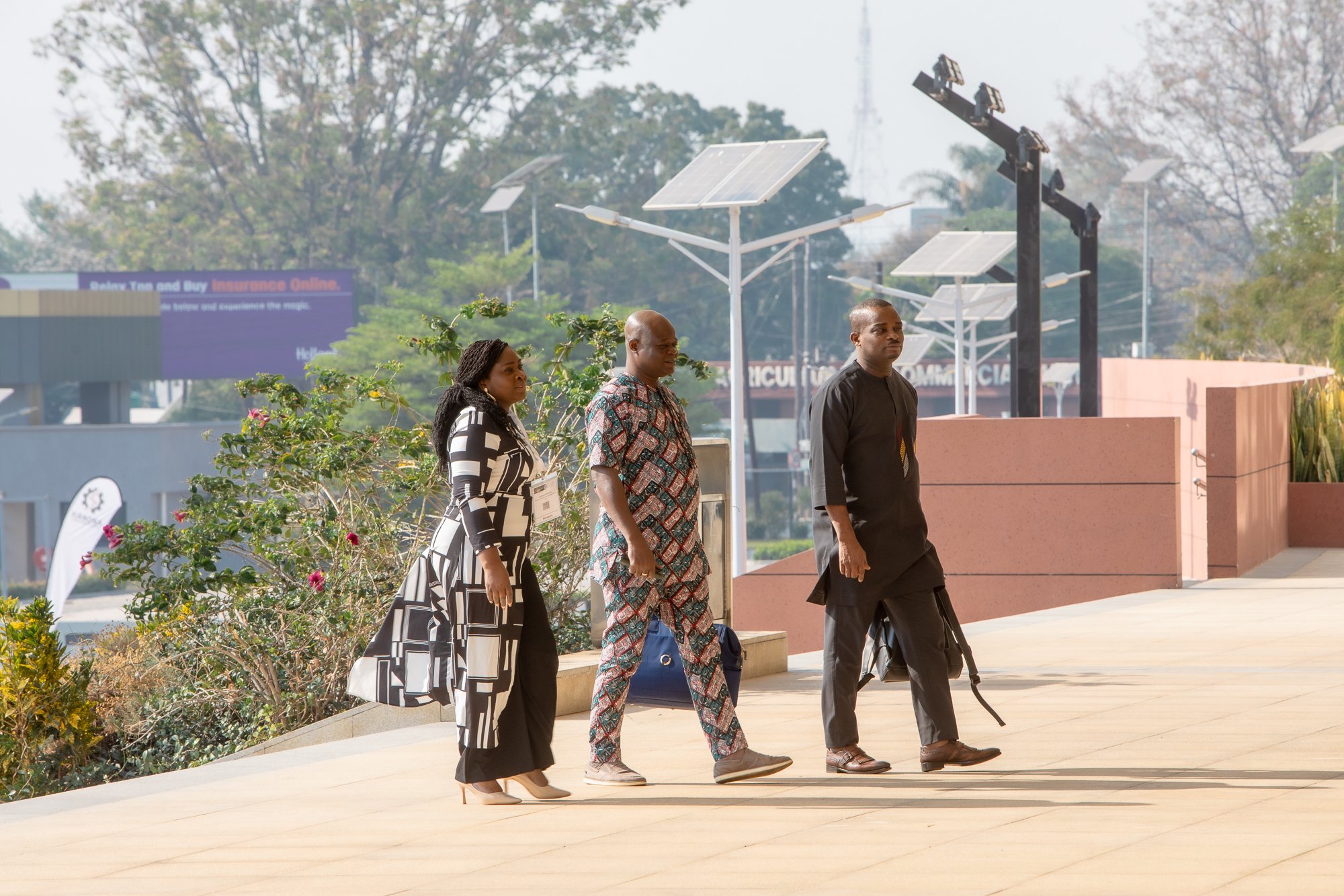 Three people walk outdoors near solar panels on a sunny day, with trees and buildings in the background. The woman on the left is dressed in a black and white patterned dress, the man in the middle wears a colorful patterned shirt, and the man on the