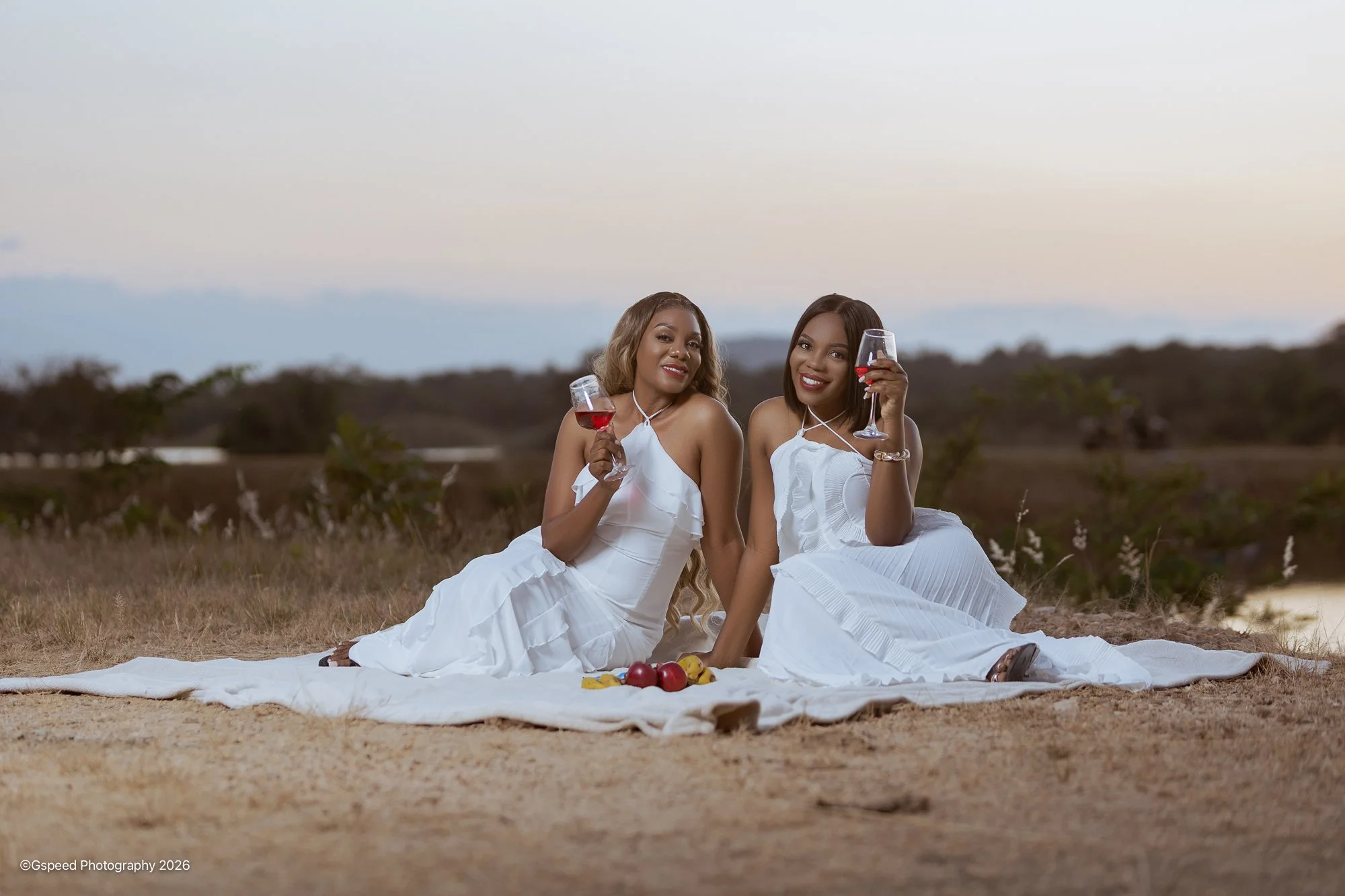 Two women sitting on a white blanket outdoors, holding glasses of red wine, with a small pile of fruits, including apples and bananas, in front of them. They are smiling and wearing white dresses, with a blurred natural landscape in the background du
