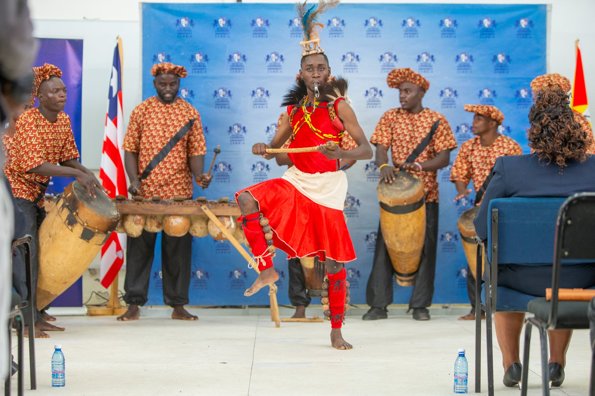 A performer in traditional African attire, with a red skirt, headdress with feathers, and face paint, dancing with a stick in front of a blue backdrop with logos, surrounded by musicians playing drums, in an indoor setting.